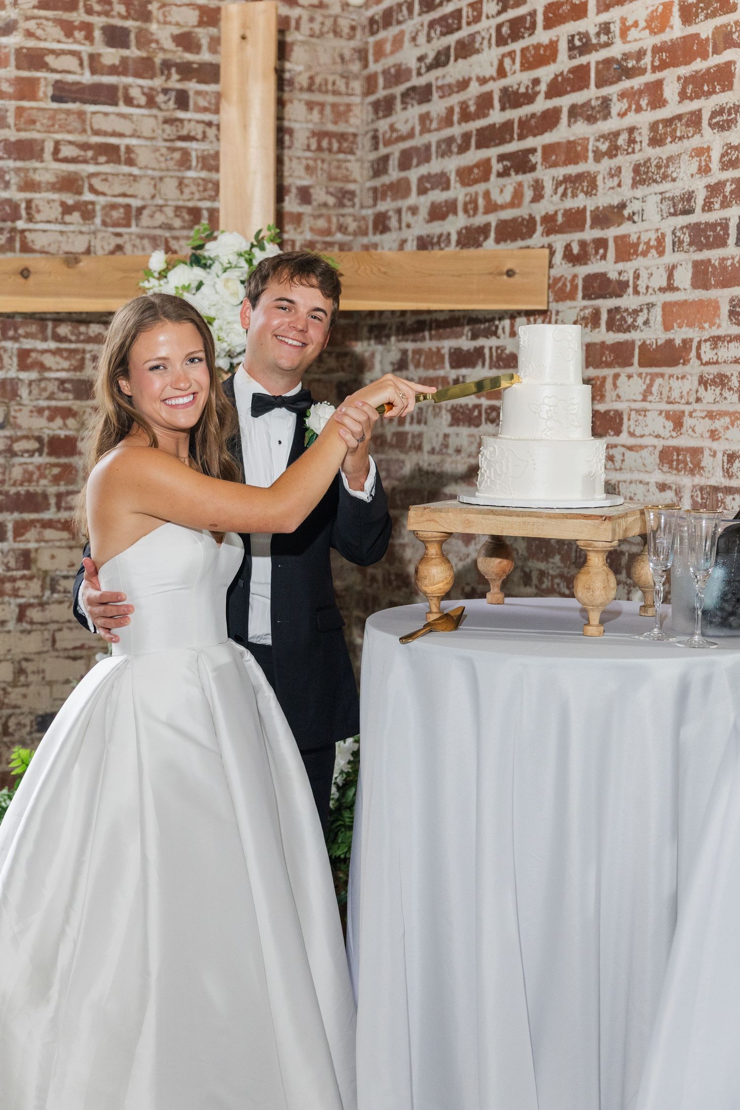 Smiling bride and groom cutting their white wedding cake at a brick-walled reception venue.