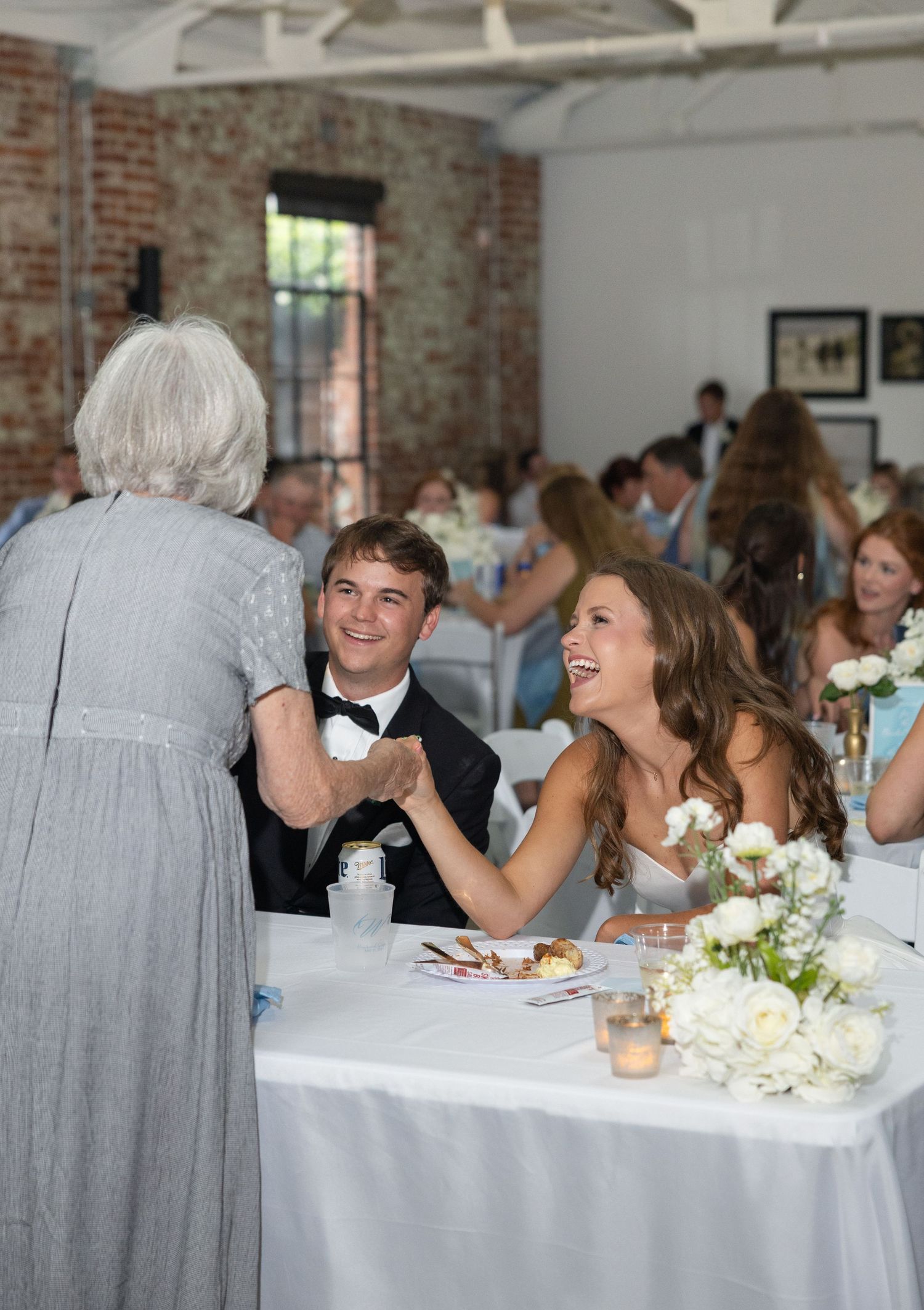 Elderly guest congratulates smiling bride and groom at decorated wedding reception table with white floral centerpieces.