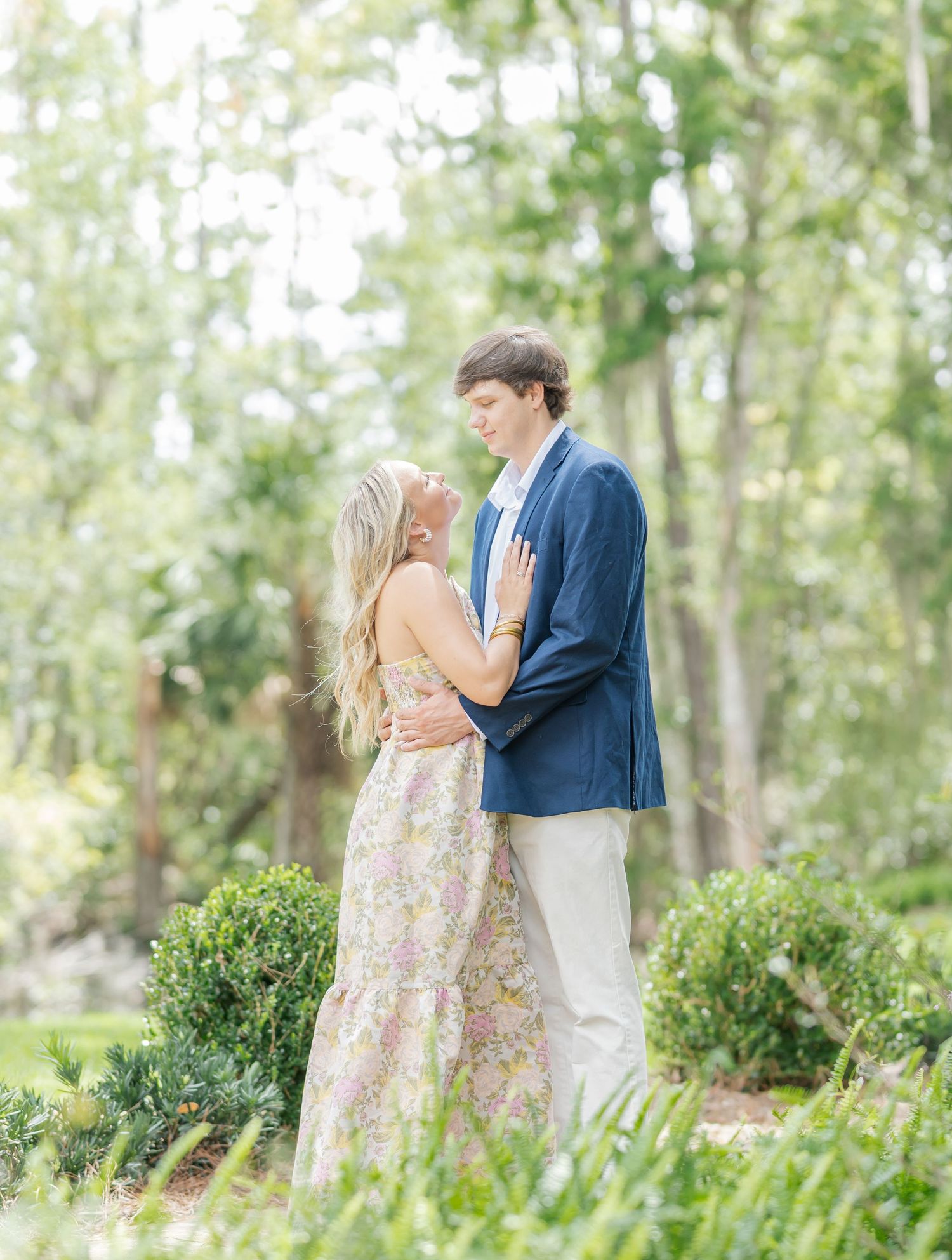 A romantic couple embraces in a garden setting with lush greenery and manicured bushes in the background.