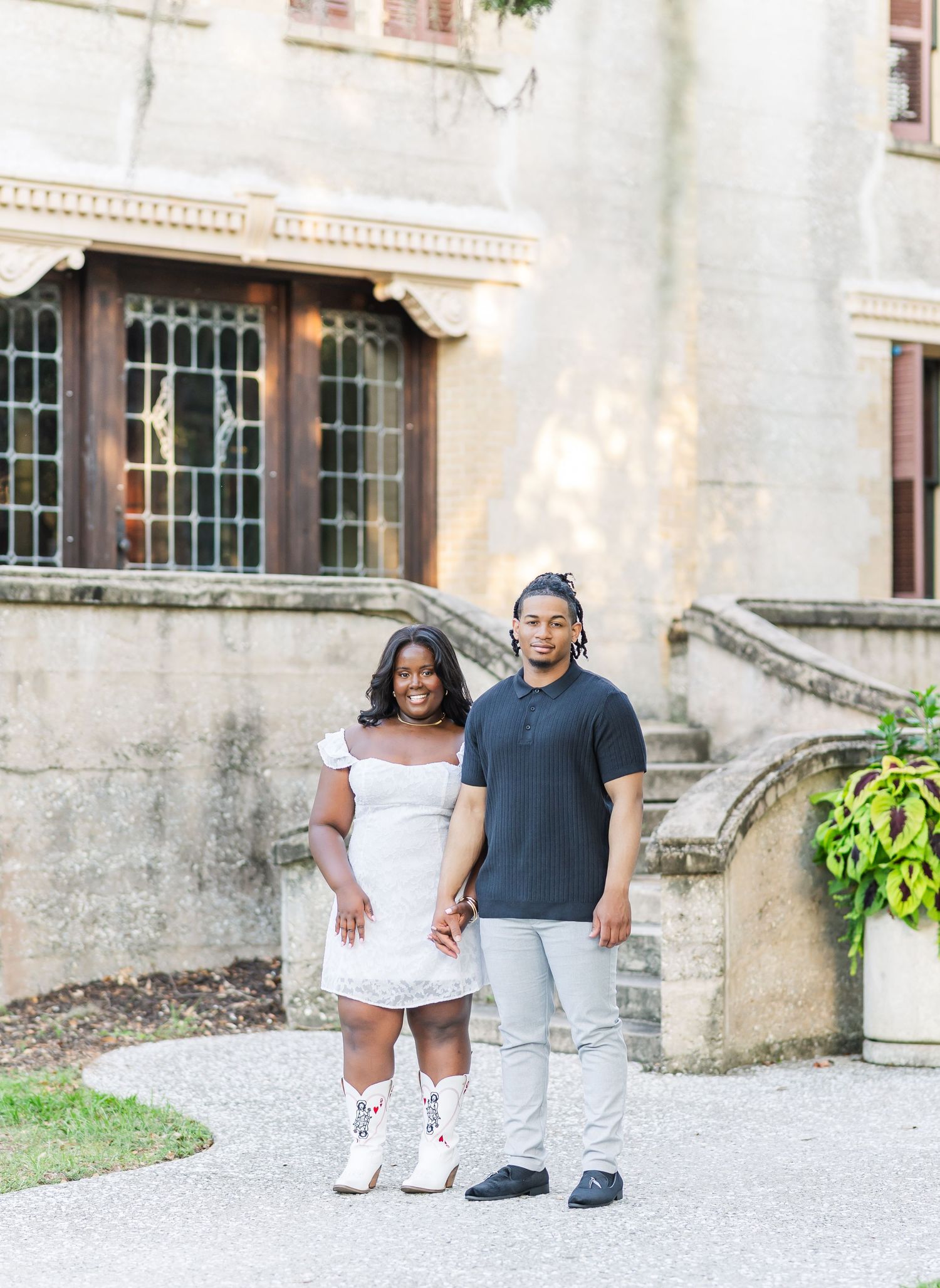 A couple walks together on a gravel path beside a historic stone building on a sunny day.