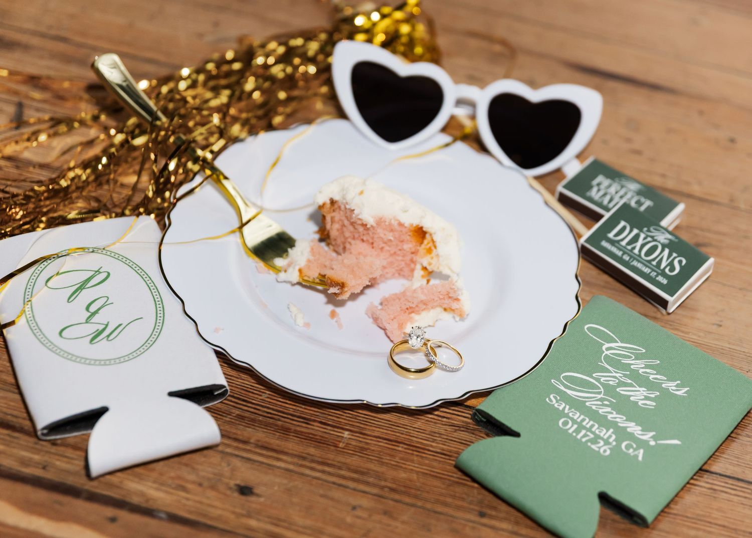 Wedding accessories including heart-shaped sunglasses, rings, and green favor koozies arranged on a wooden surface.