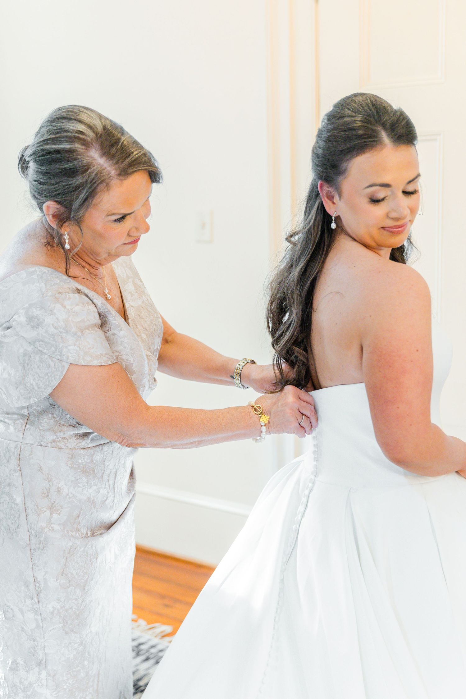 Someone in a white lace robe helps button up a white wedding dress during bridal preparations.