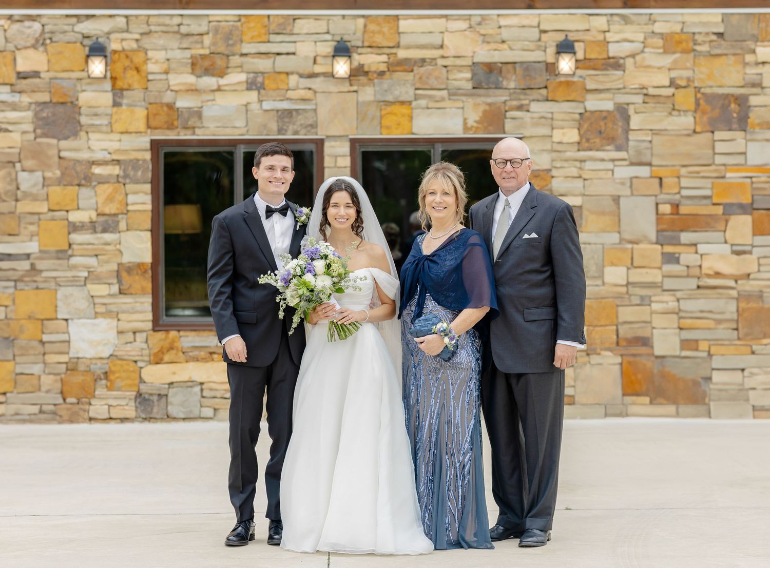 Wedding party poses in front of rustic stone wall with elegant attire and floral bouquet.