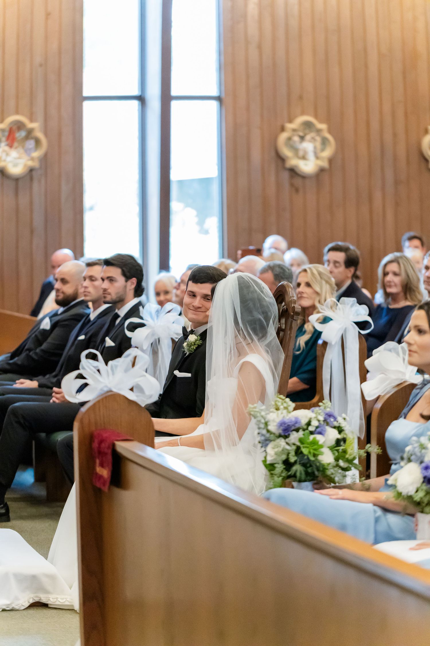 Wedding guests seated in wooden church pews decorated with white bows.