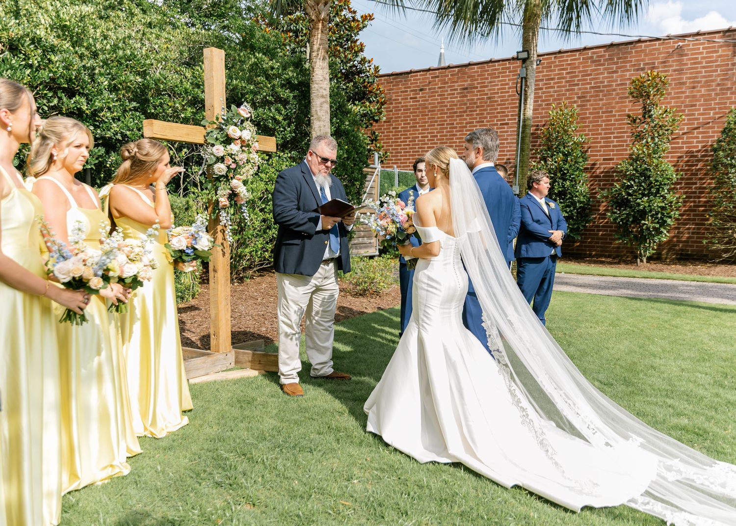 An outdoor wedding ceremony takes place in front of a wooden cross with bridesmaids in yellow dresses standing to the side.