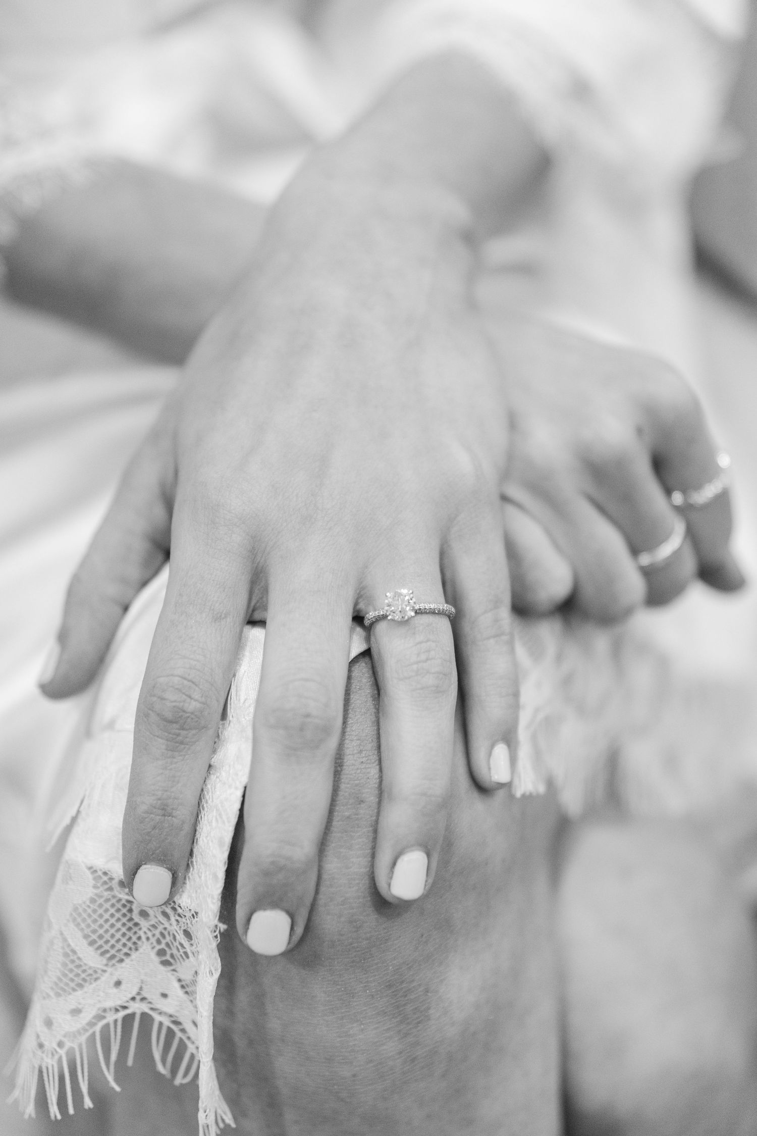 Black and white close-up photo of hands showing an engagement ring with lace detail in the background.