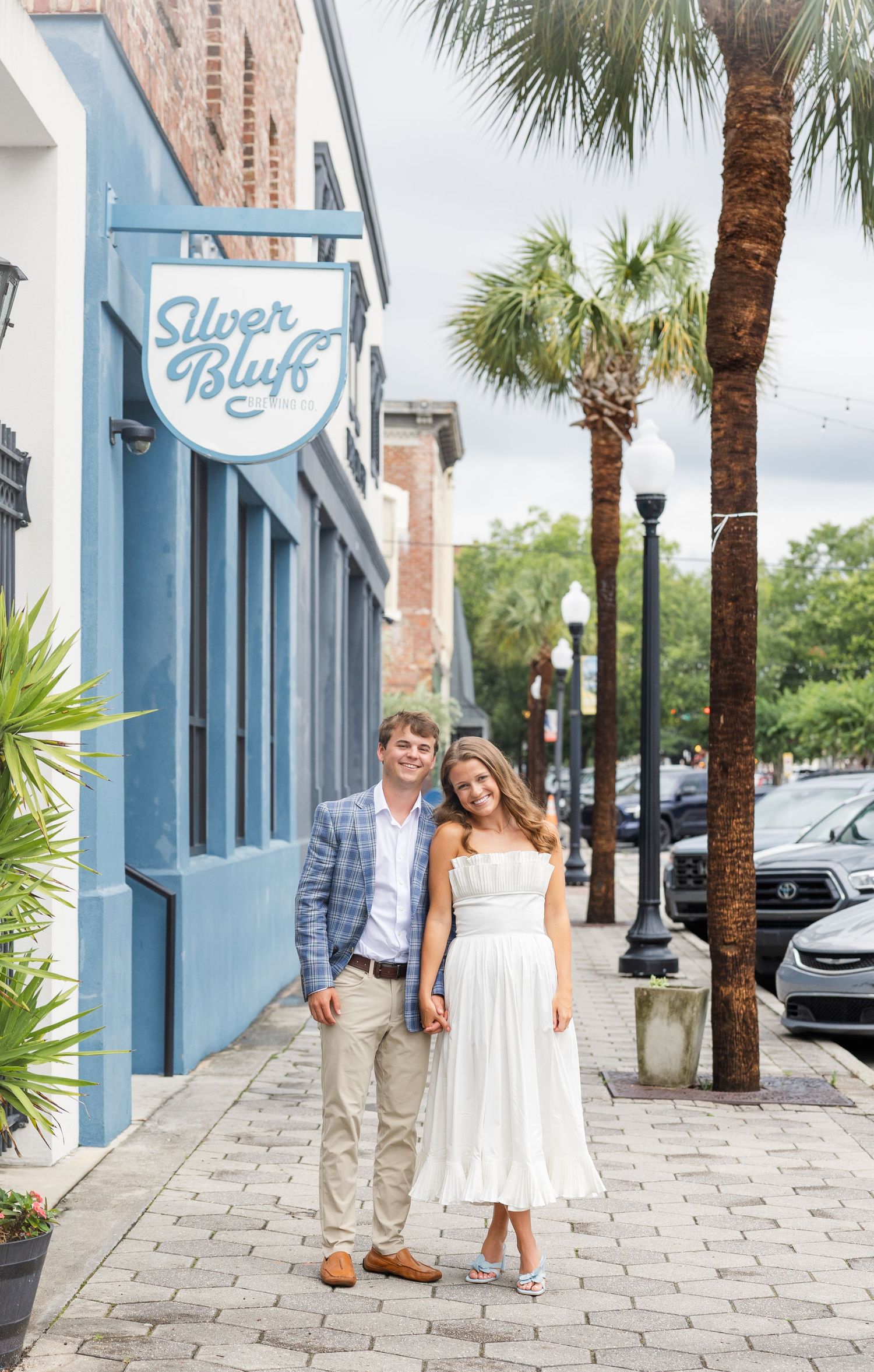 Couple posing outside Silver Bluff brewery on a palm-lined sidewalk, woman in white dress.