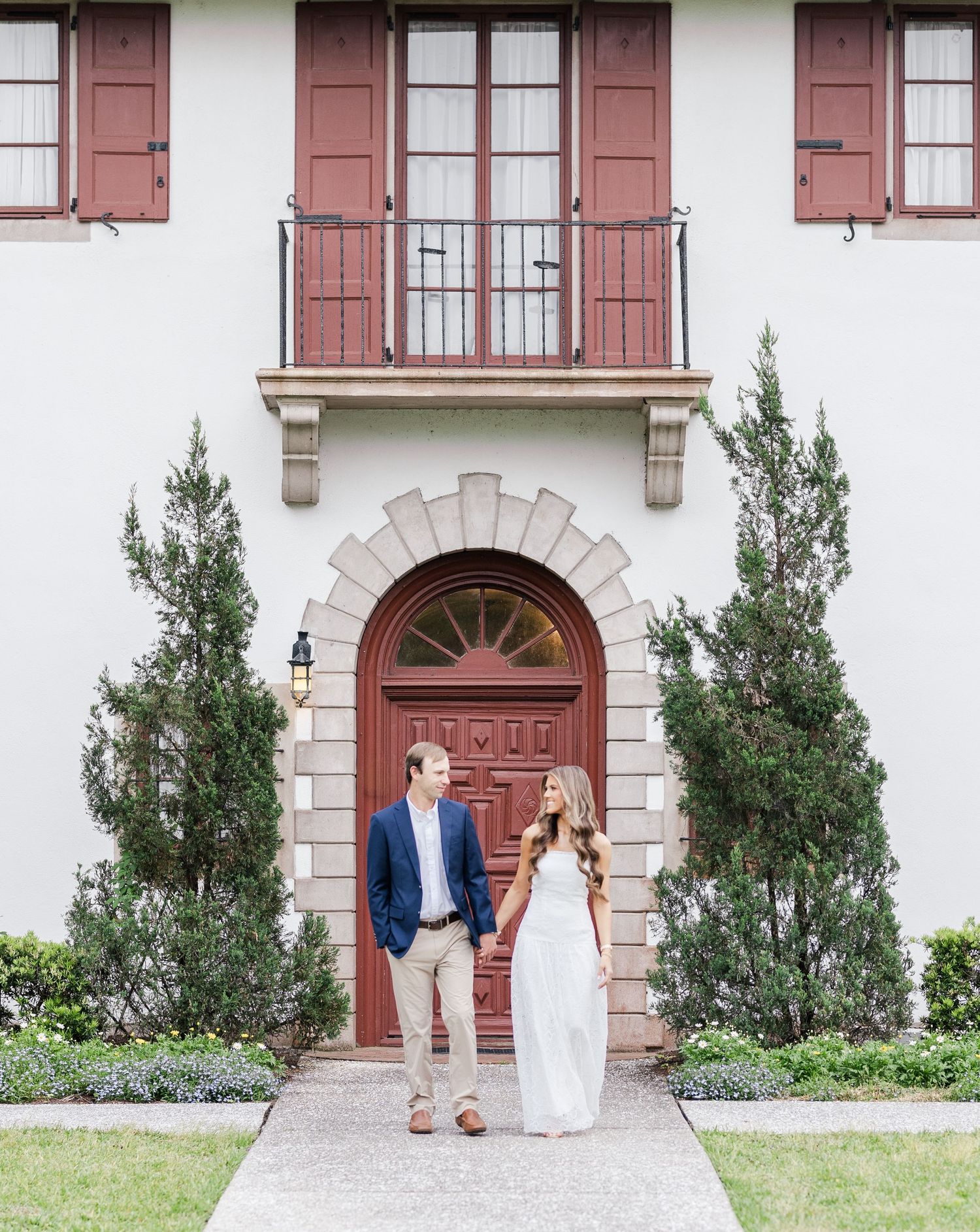 Couple holding hands in front of elegant red arched door with cypress trees at a Mediterranean-style estate.