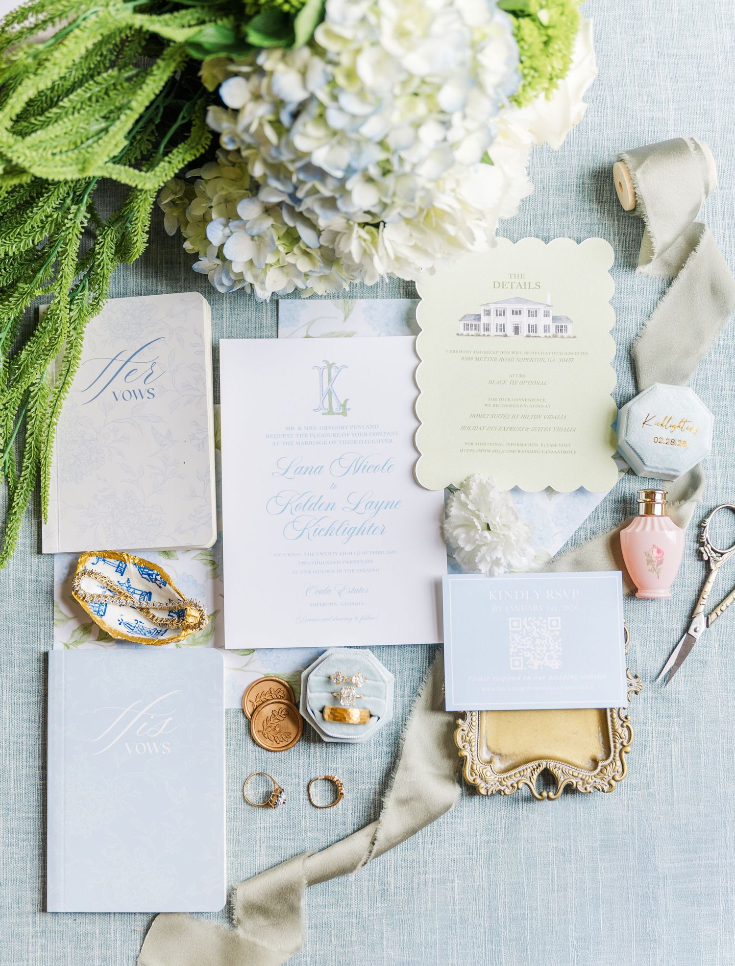 Elegant wedding invitation suite with blue stationery, gold rings, pearl jewelry, floral accents, and ribbon on a soft blue background.