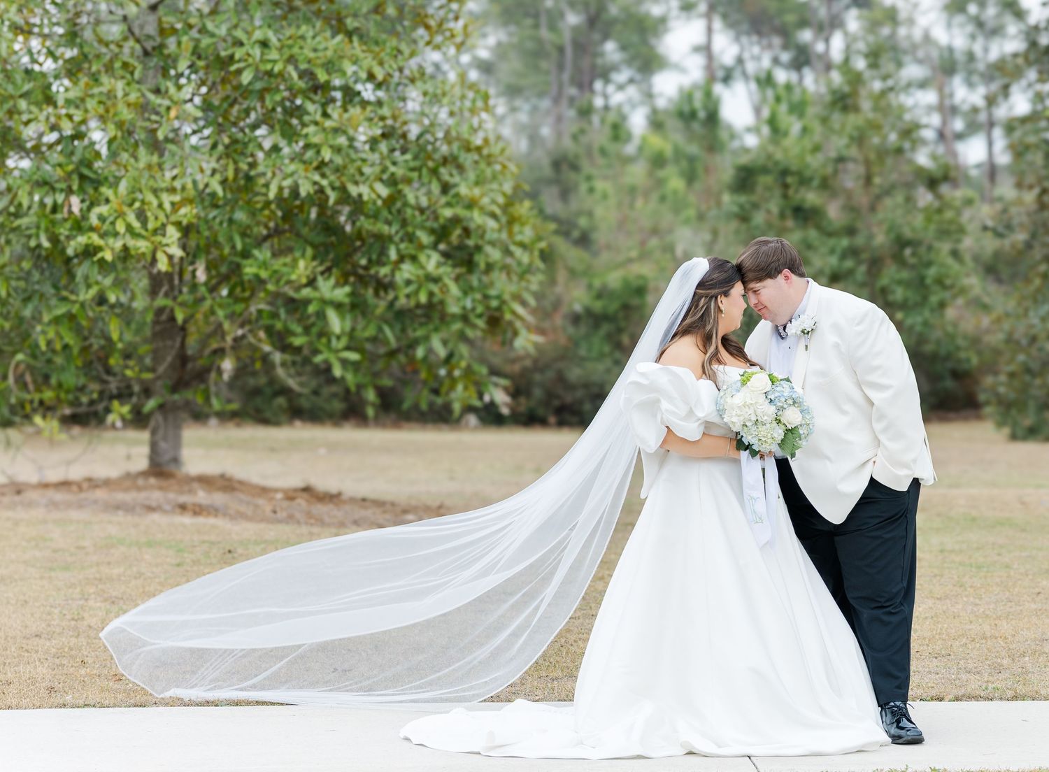 Bride and groom sharing a romantic kiss outdoors, her cathedral veil flowing in the wind against a lush green backdrop.