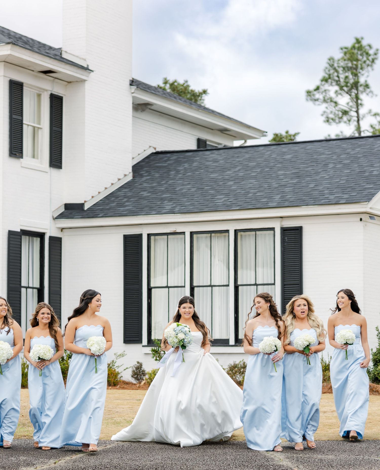 Bride in white ball gown walks with bridesmaids in light blue strapless gowns outside a white wedding venue.