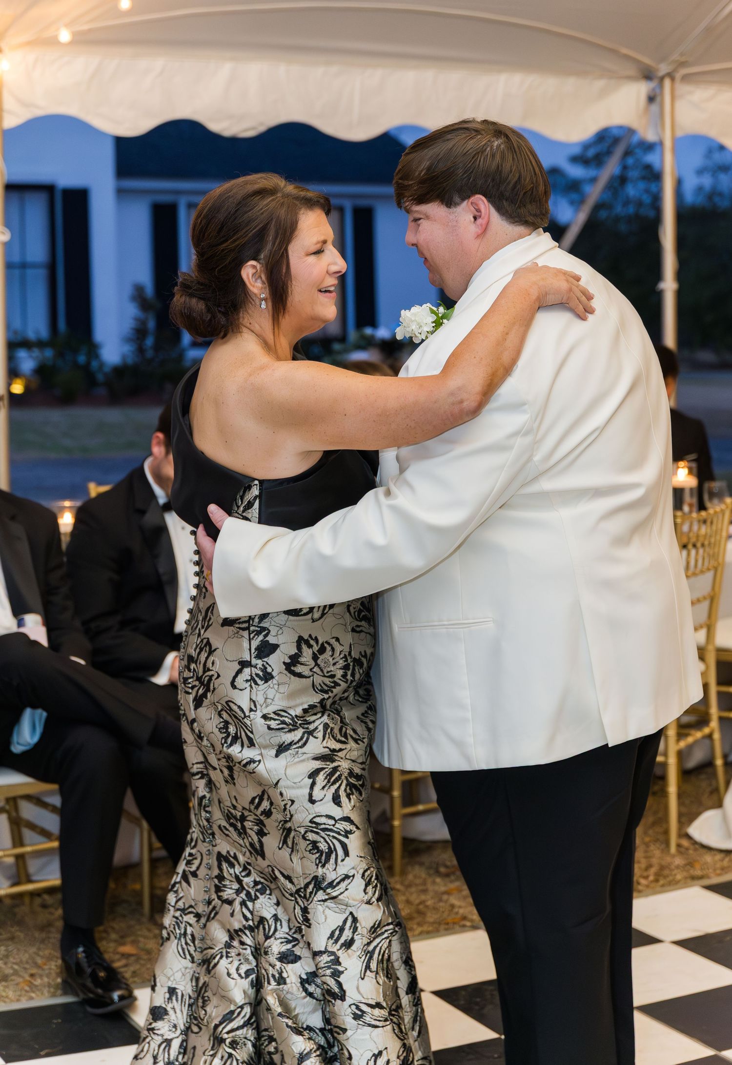 Groom in white tuxedo jacket shares mother-son dance under tent at outdoor wedding reception.