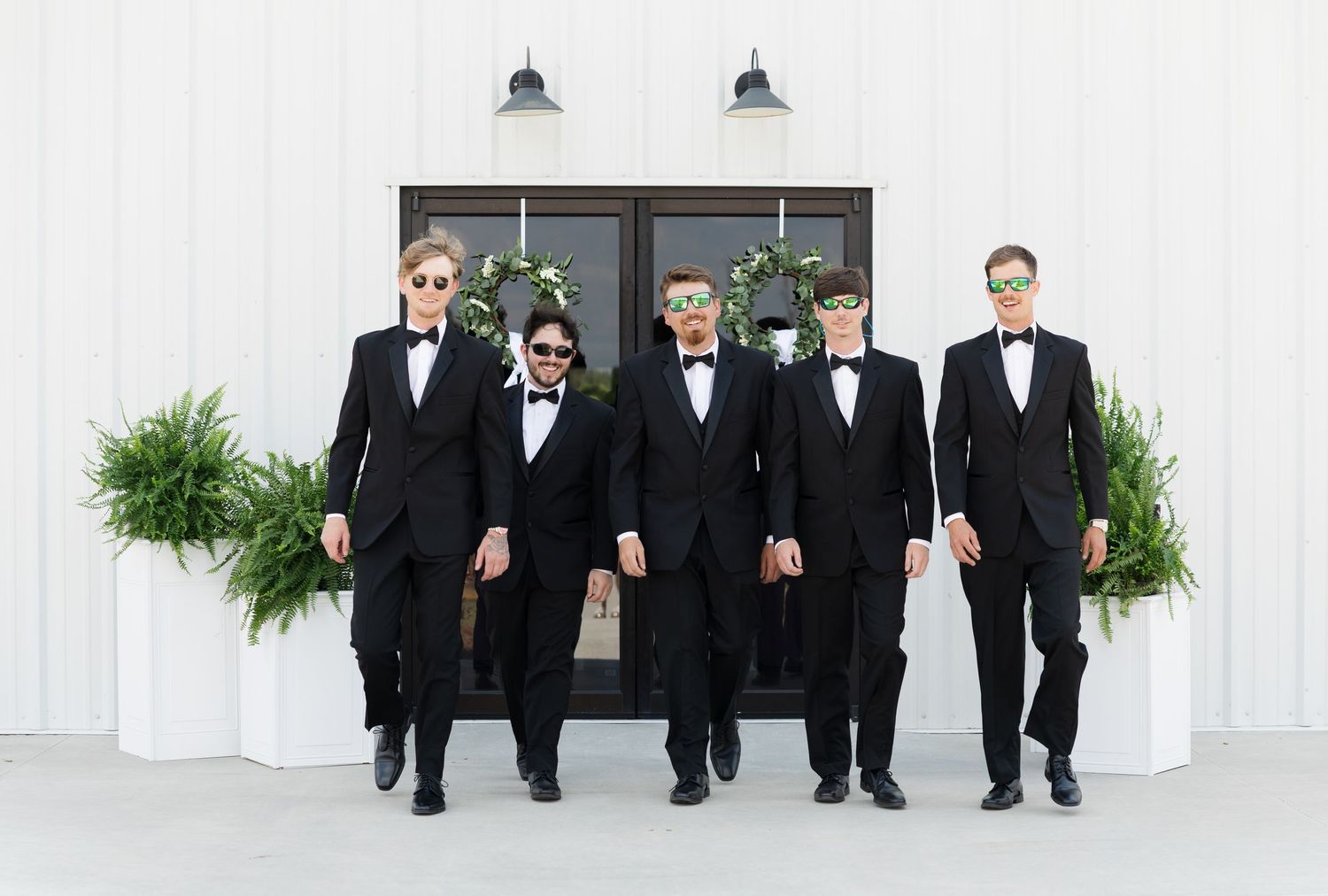 Five groomsmen in black tuxedos and bow ties pose confidently outside a white barn venue.