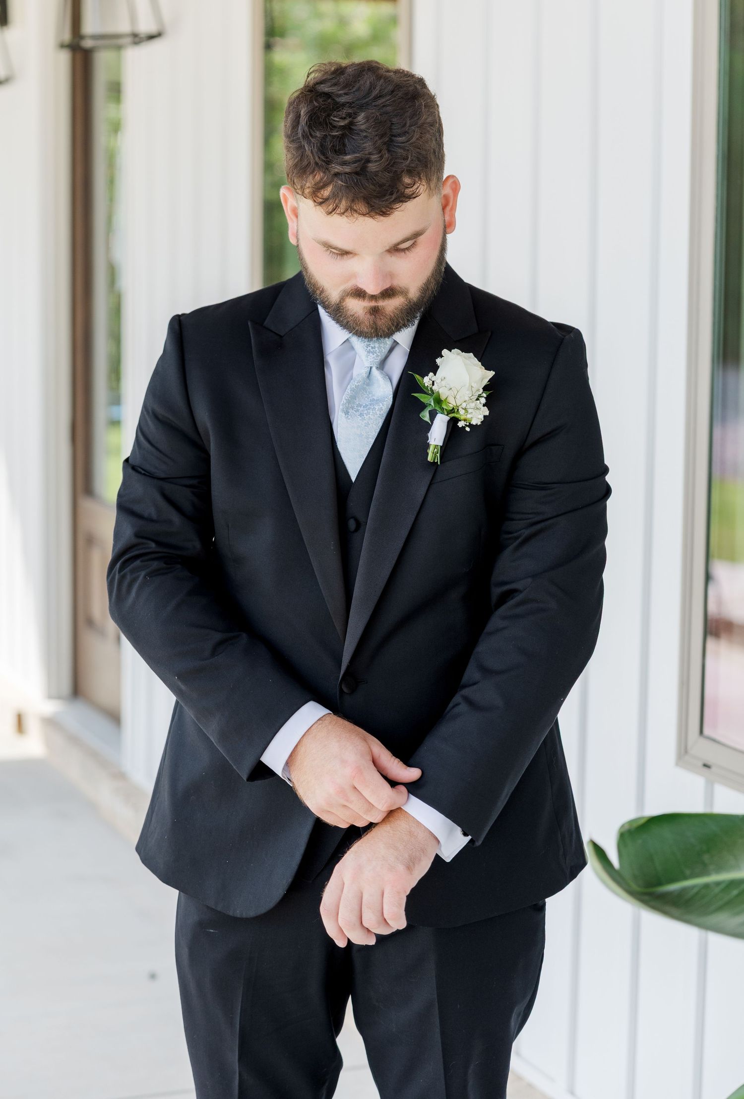 Groom in black tuxedo with light blue tie and white boutonniere adjusting his cufflinks before wedding.