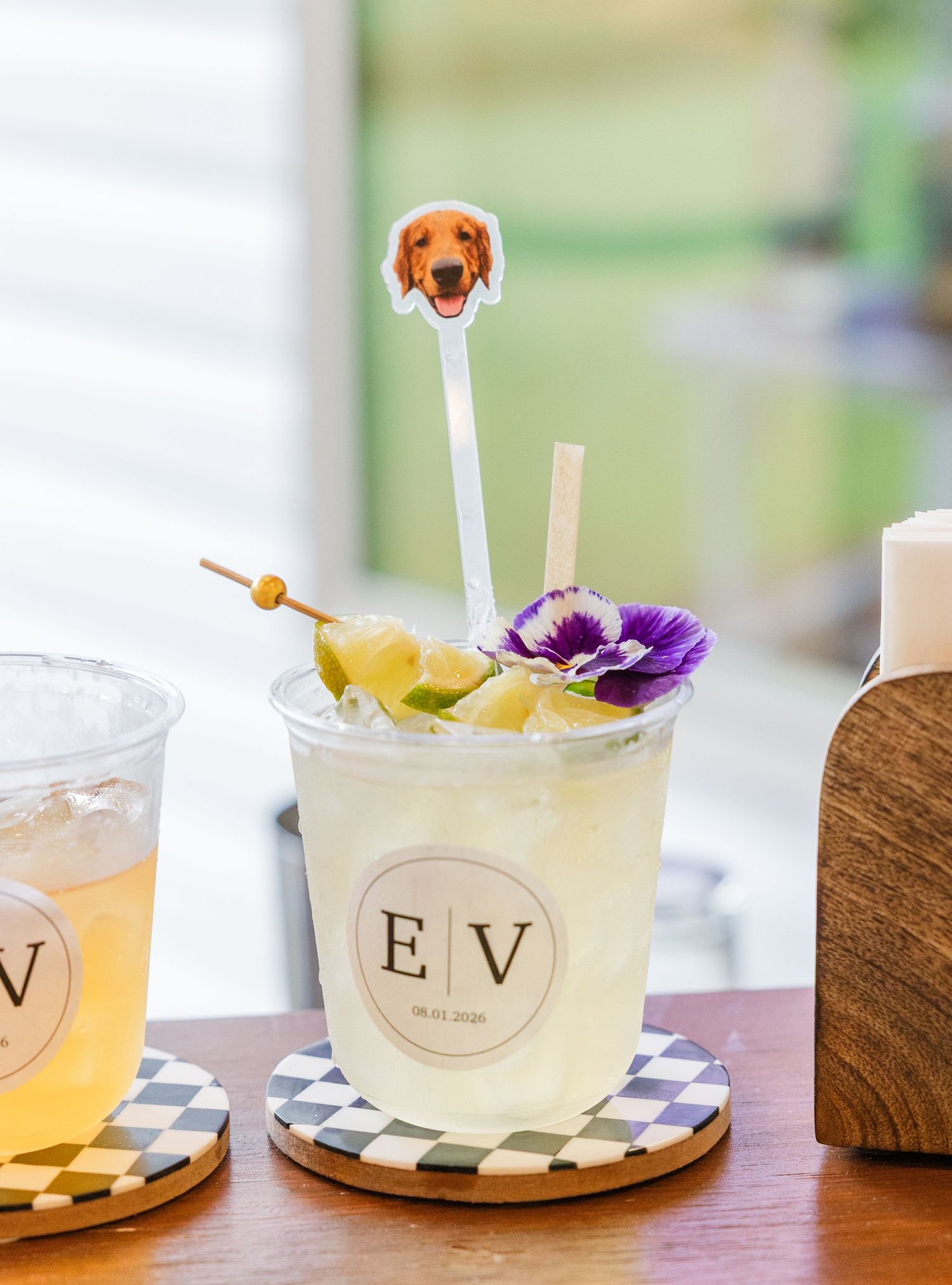 A cocktail in an EV monogram cup with a dog stir stick, purple flower, and fruit garnish on a checkered coaster.
