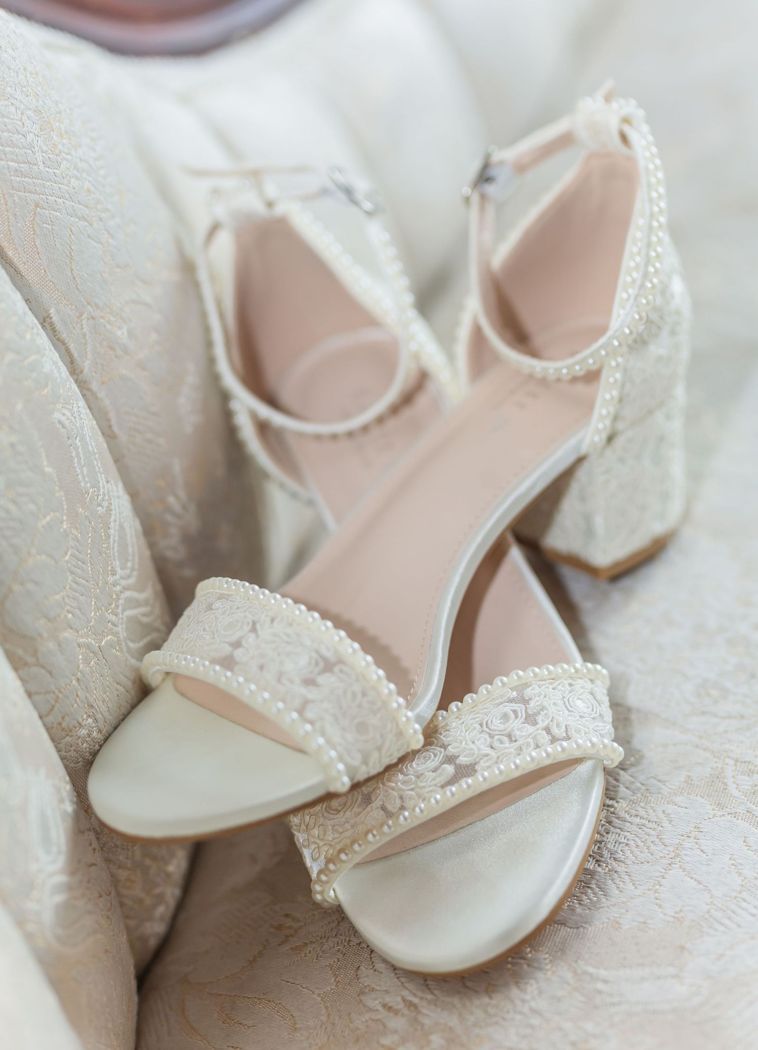Delicate white lace bridal shoes with pearl details displayed on white fabric.