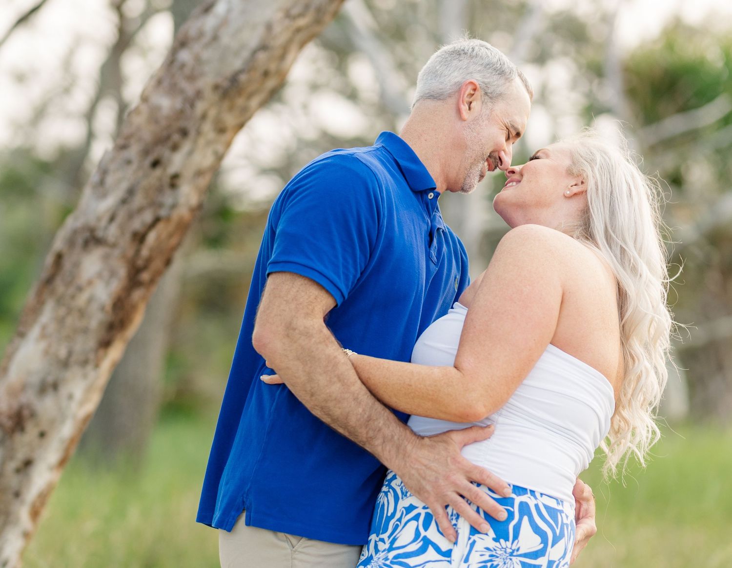 A romantic couple shares an intimate moment outdoors near a tree, with the person in blue embracing their partner.