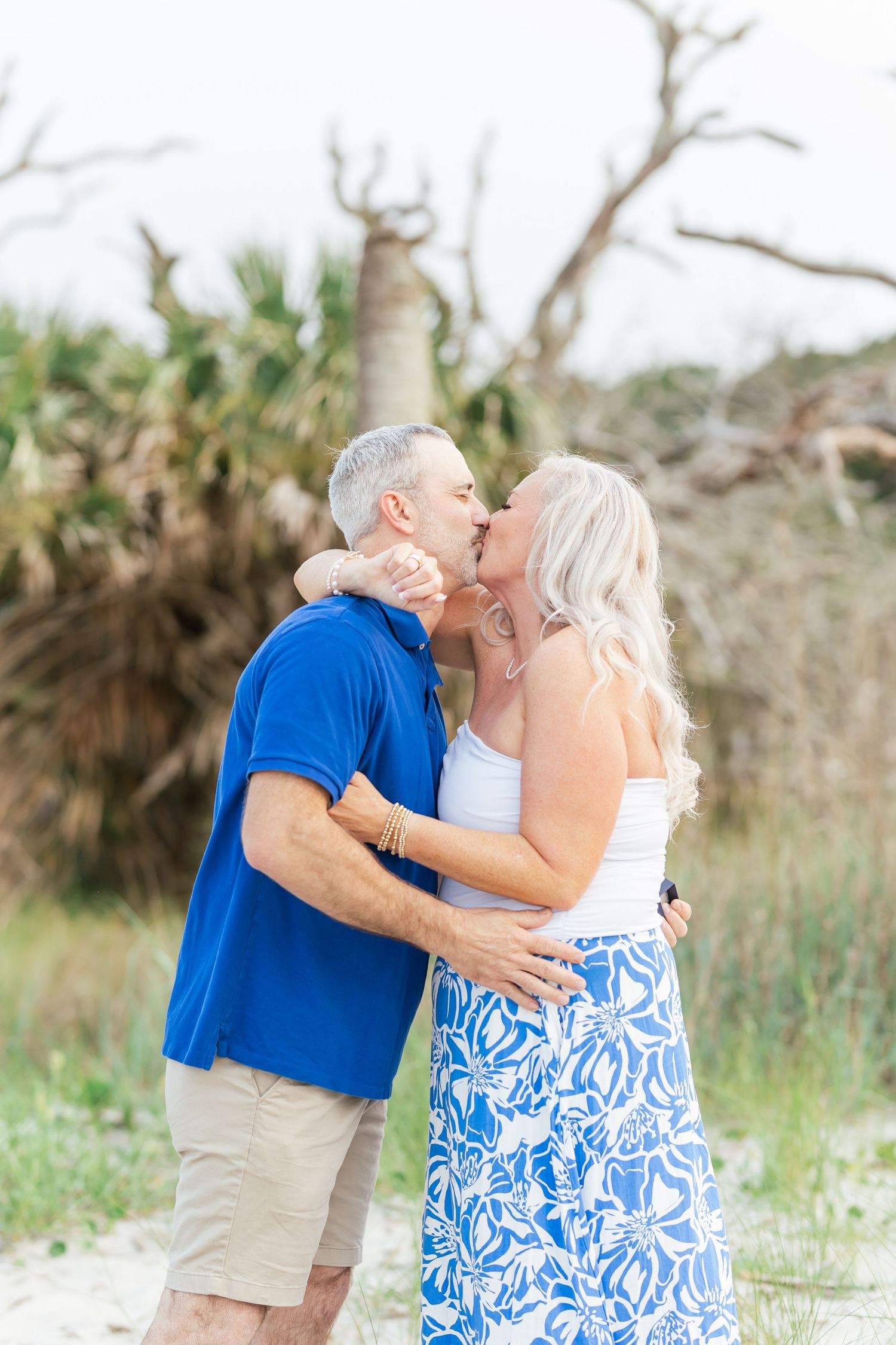 A romantic beach engagement kiss surrounded by natural coastal vegetation.