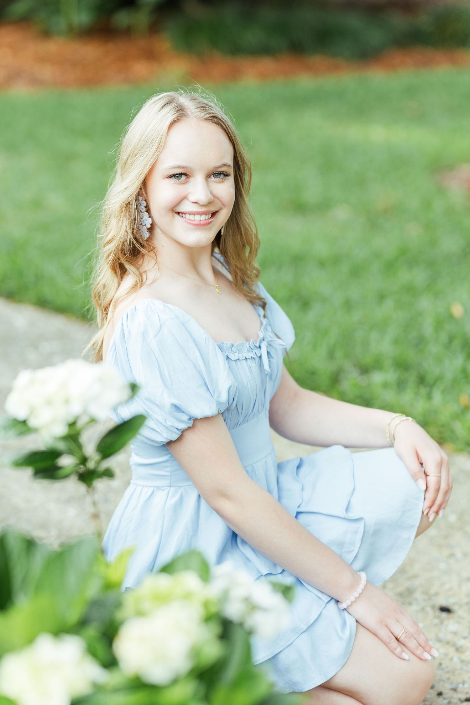 A person in a light blue summer dress sits gracefully on stone steps surrounded by white hydrangea flowers in a garden.