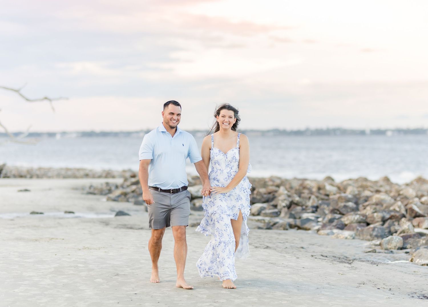 A couple walks hand in hand on a rocky beach at sunset with a waterfront view in the background.