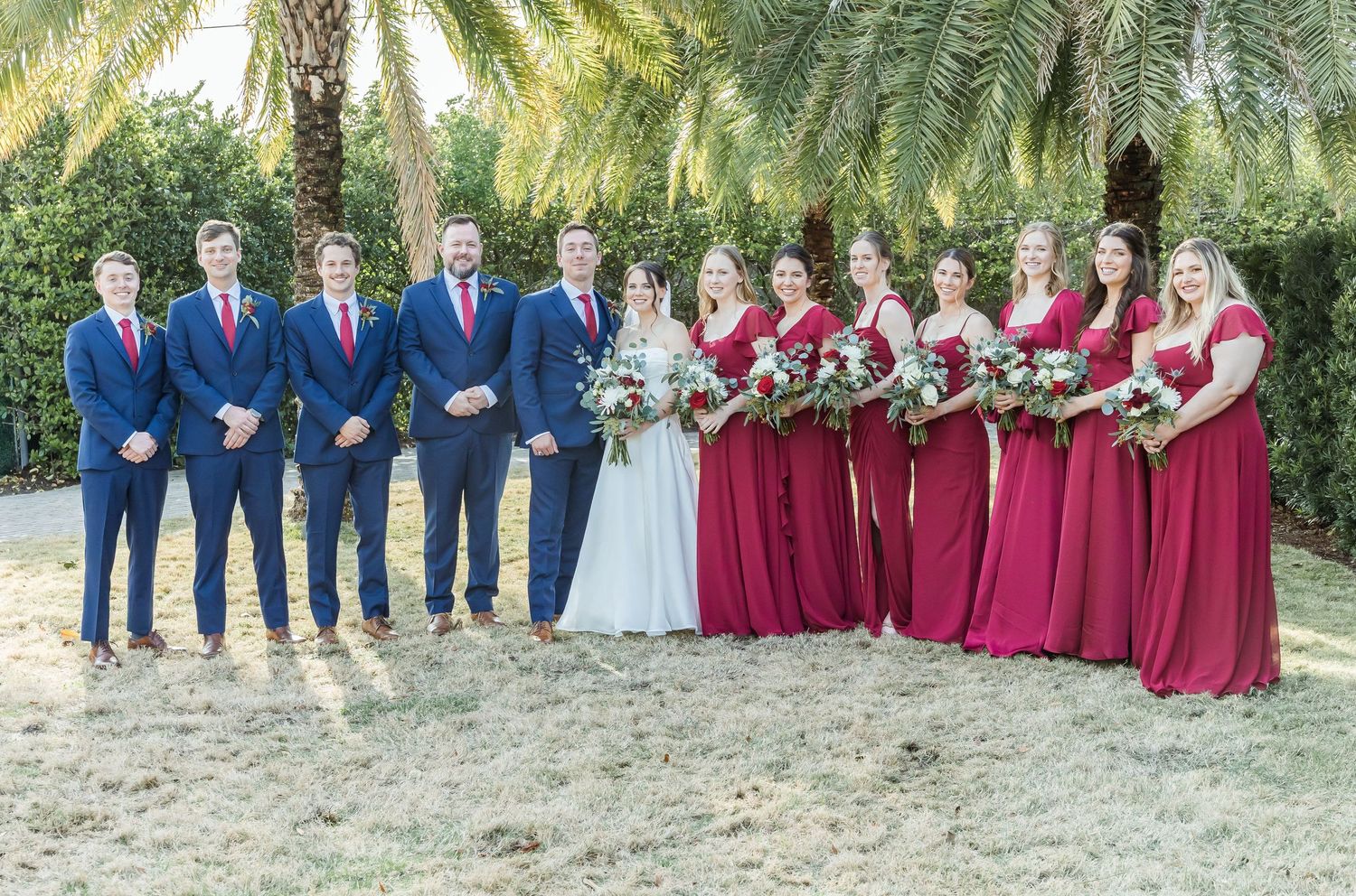 Wedding party lineup under palm trees with burgundy dresses and navy suits.
