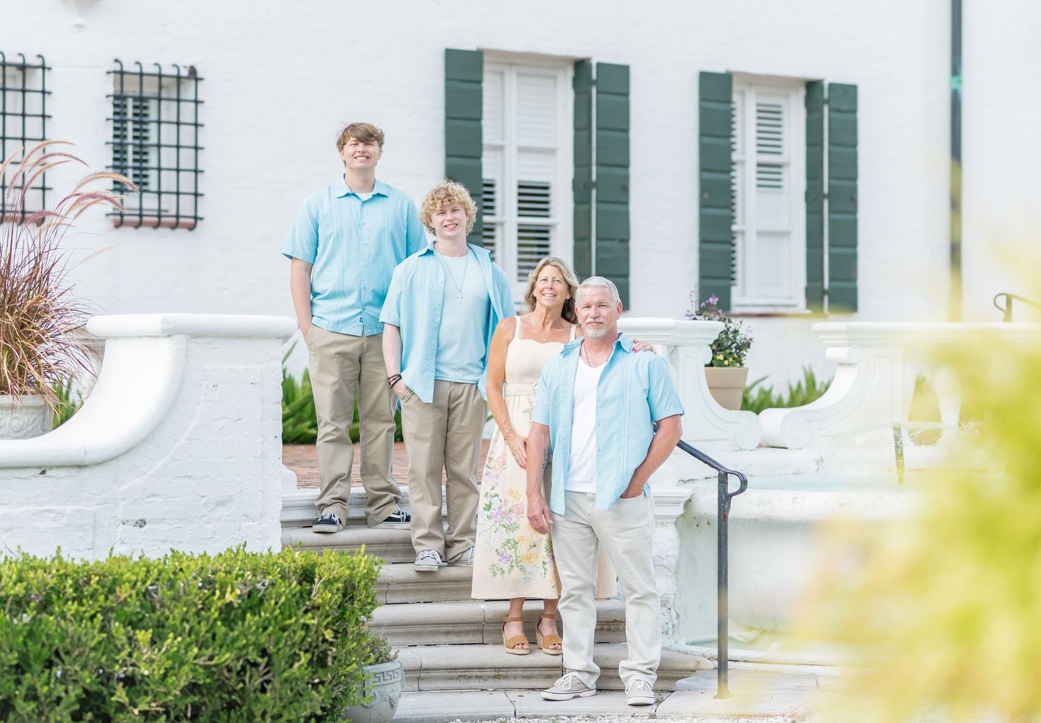 Group in coordinating blue shirts and khaki pants standing on exterior white steps of a house with green shutters.