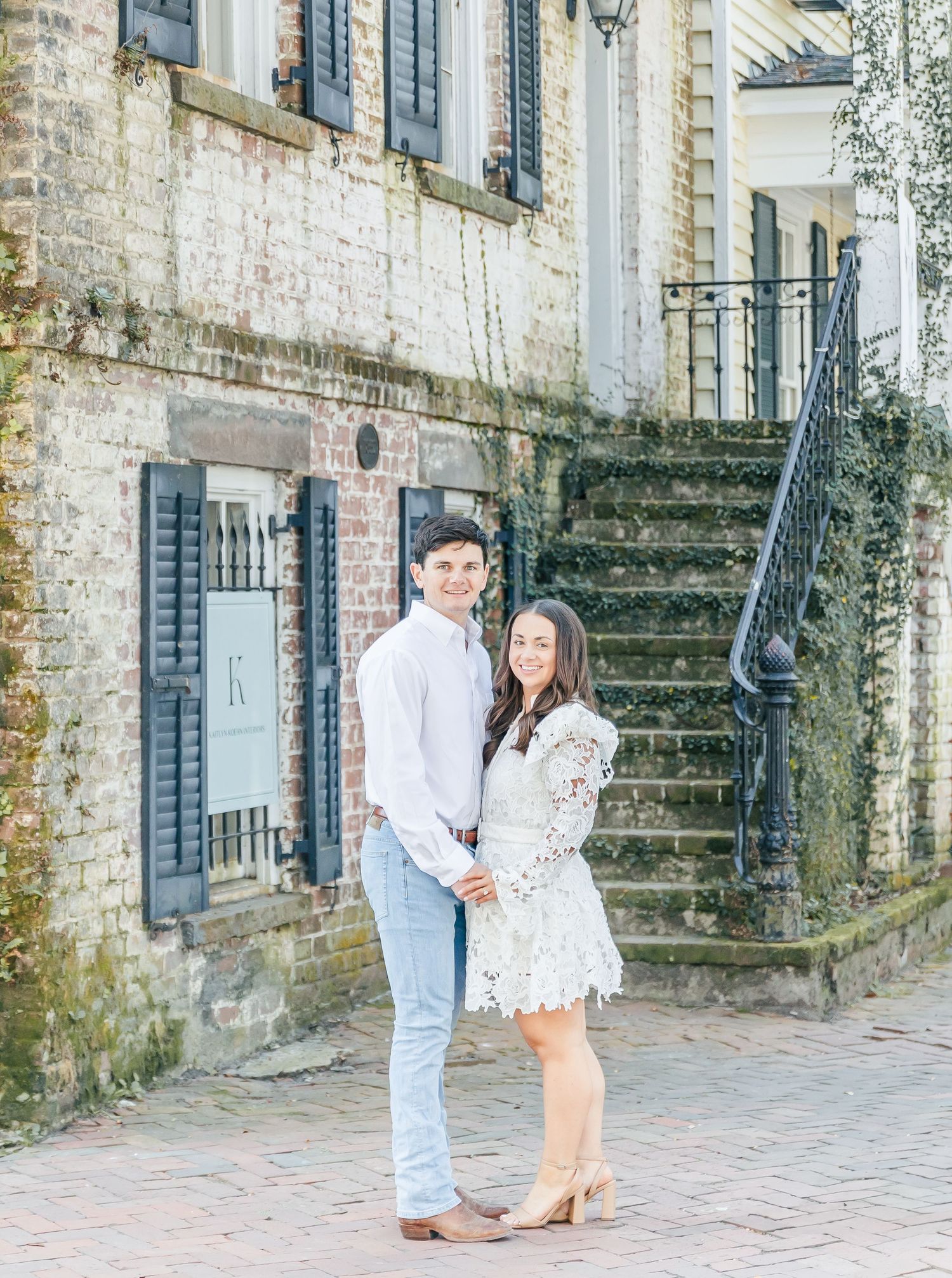 A couple poses on cobblestone street beside weathered brick walls and iron railing staircase.