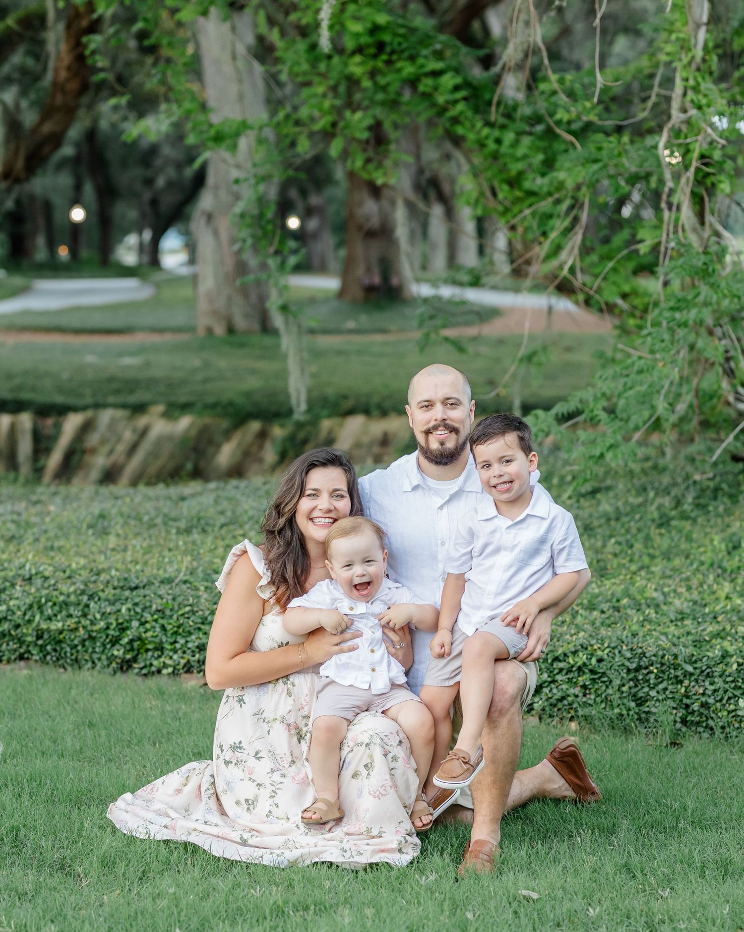 A family sits together on a white blanket during an outdoor photo session in a lush green park setting.