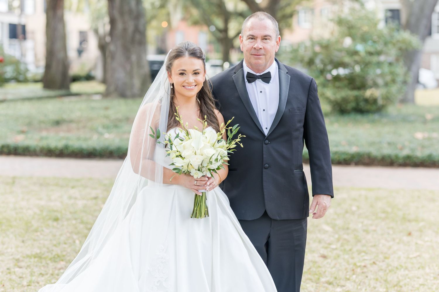 Bride in white gown holding bouquet walks with tuxedo-clad groom outdoors at elegant wedding ceremony.