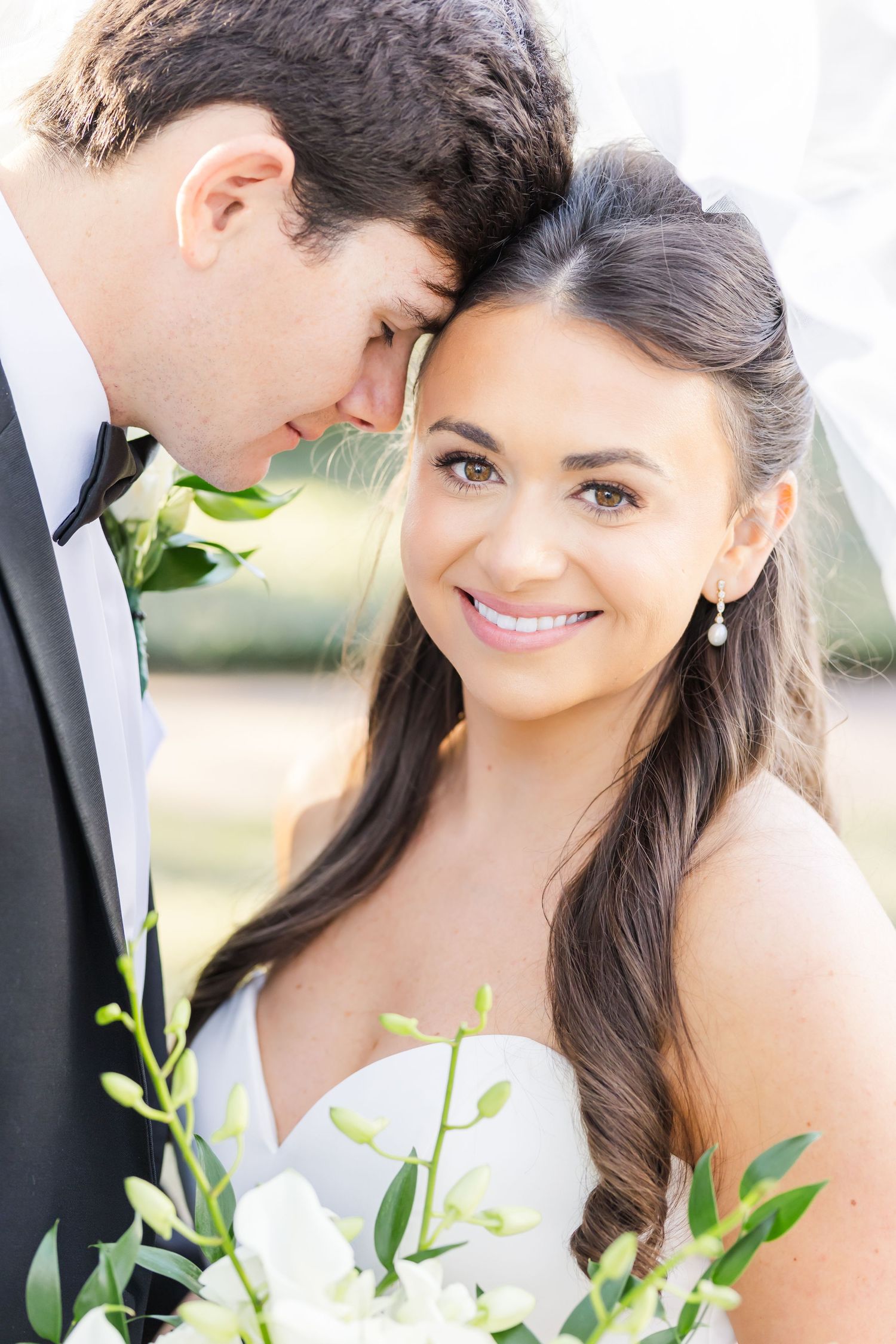 Close-up portrait of smiling bride with dark hair holding white bouquet as groom leans in tenderly.