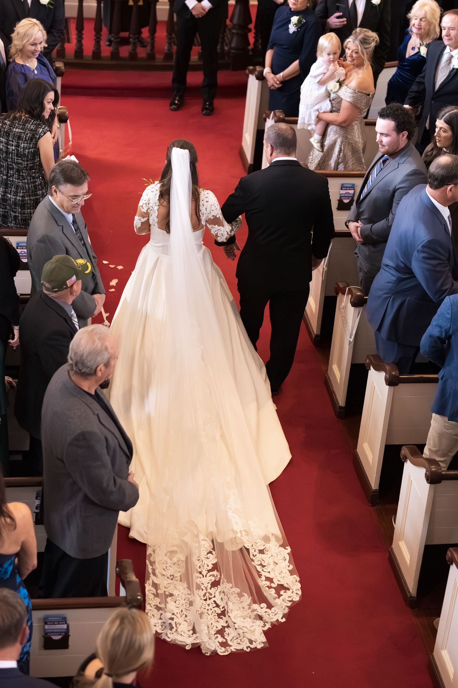 Bride in lace-trimmed gown and veil walking down red carpet aisle with escort.