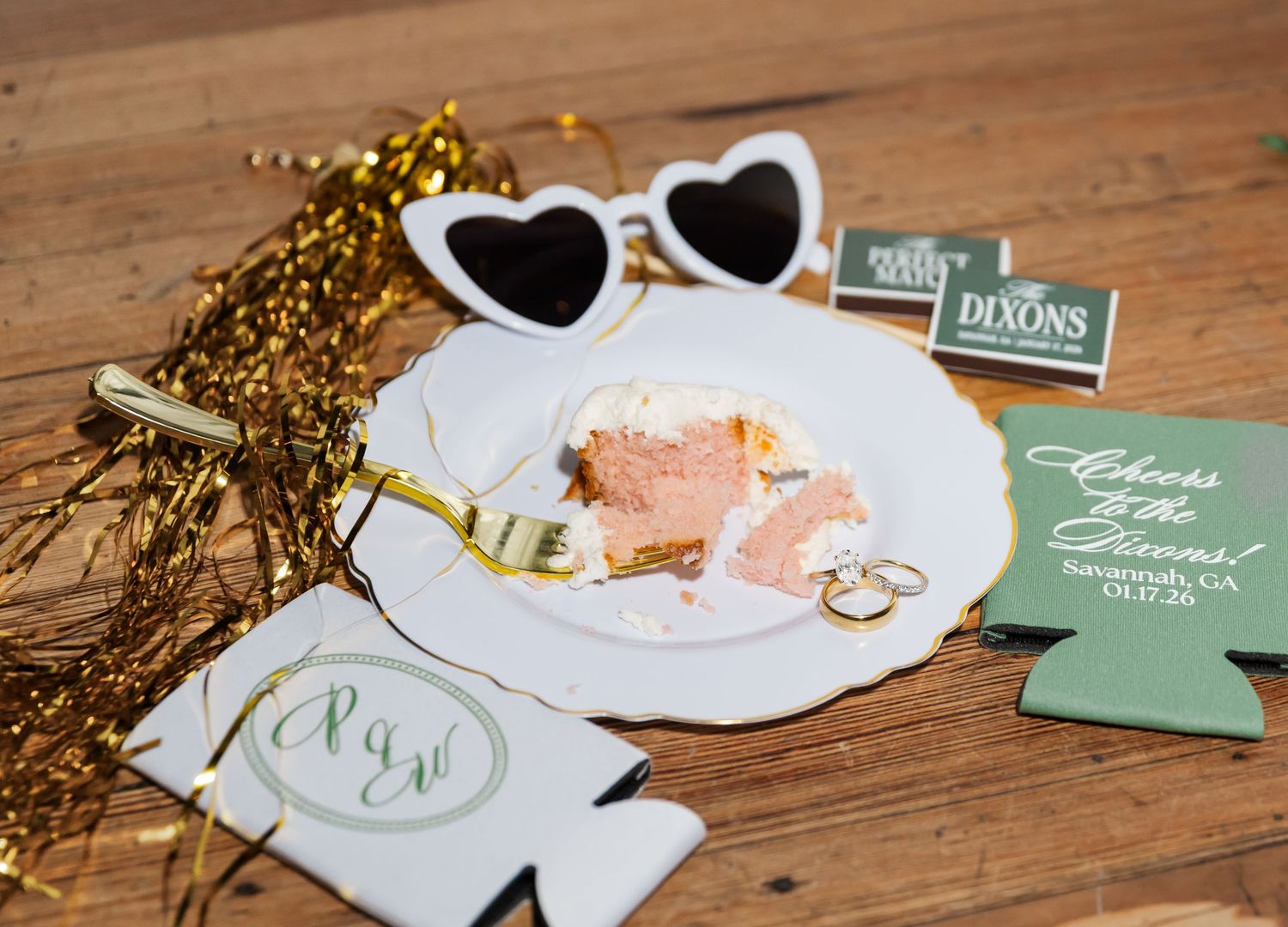 Wedding flat lay with heart sunglasses, cake slice, gold rings, koozies, and sparkly decorations on wood.