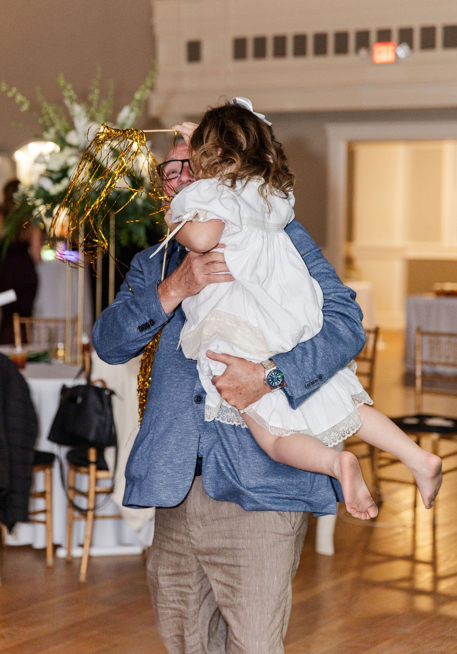 Man in blue blazer lifting barefoot flower girl on wedding reception dance floor.