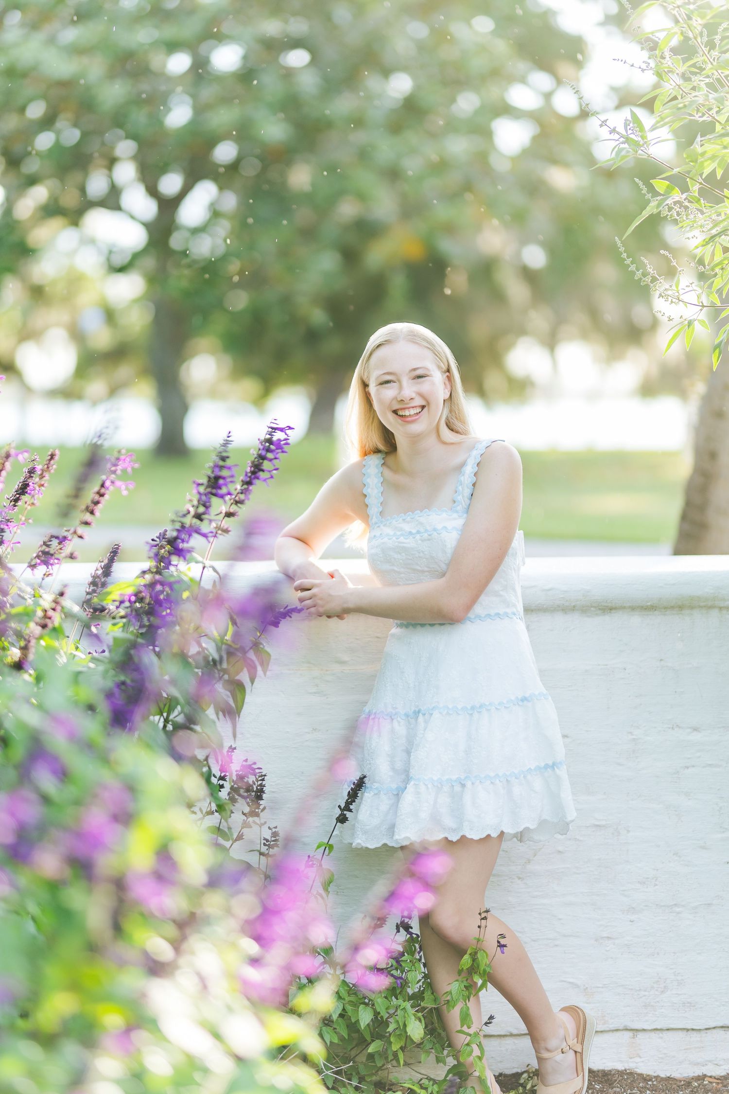 A white eyelet lace dress twirls gracefully in a garden setting with purple flowers in the foreground.