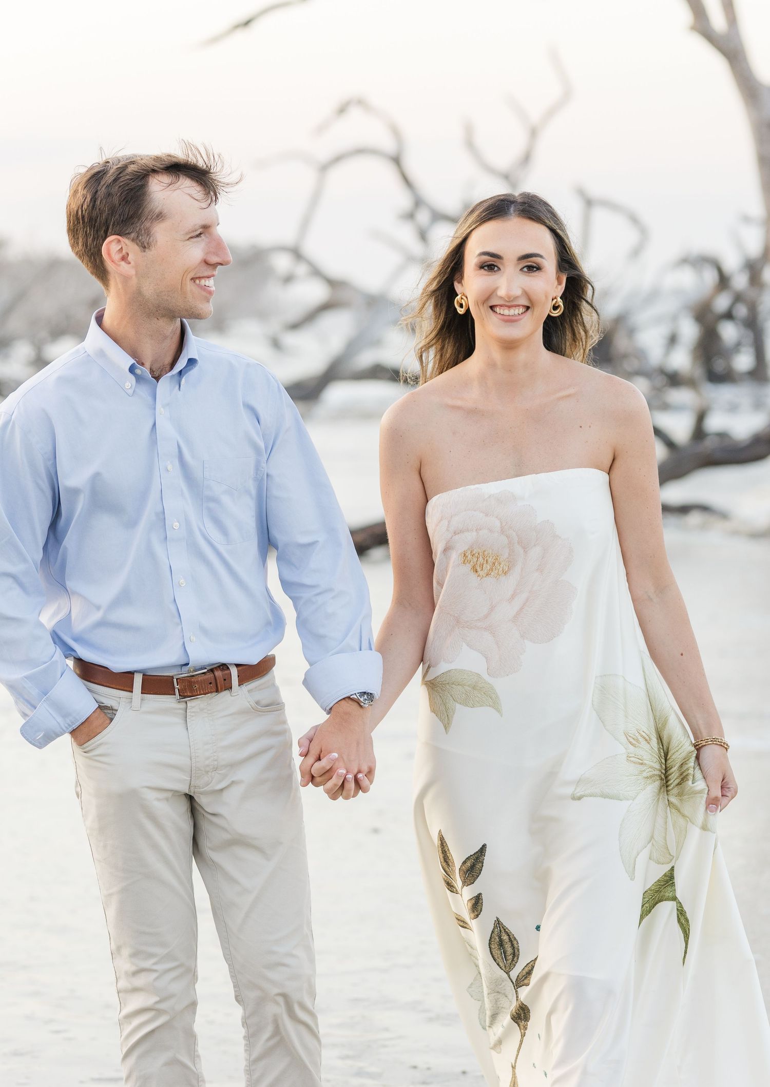 Smiling couple strolling hand in hand on sandy beach, woman in elegant floral dress, man in blue button-down shirt.
