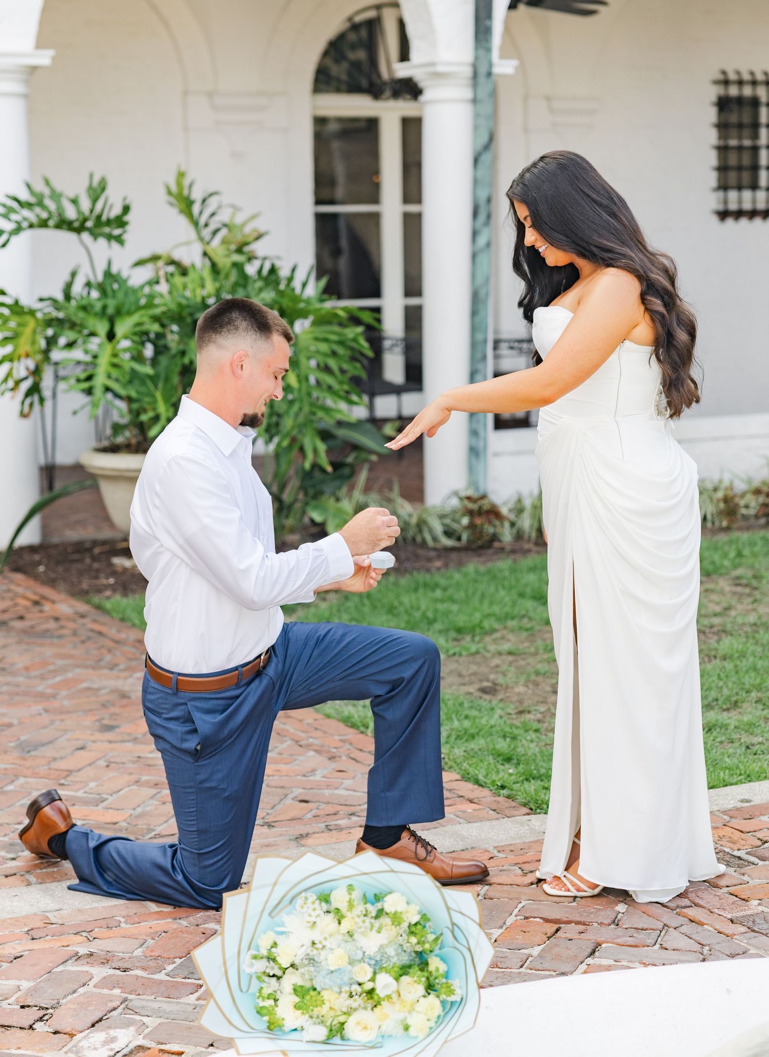 Man kneeling on brick patio proposing to woman in white dress with bouquet of white flowers nearby.