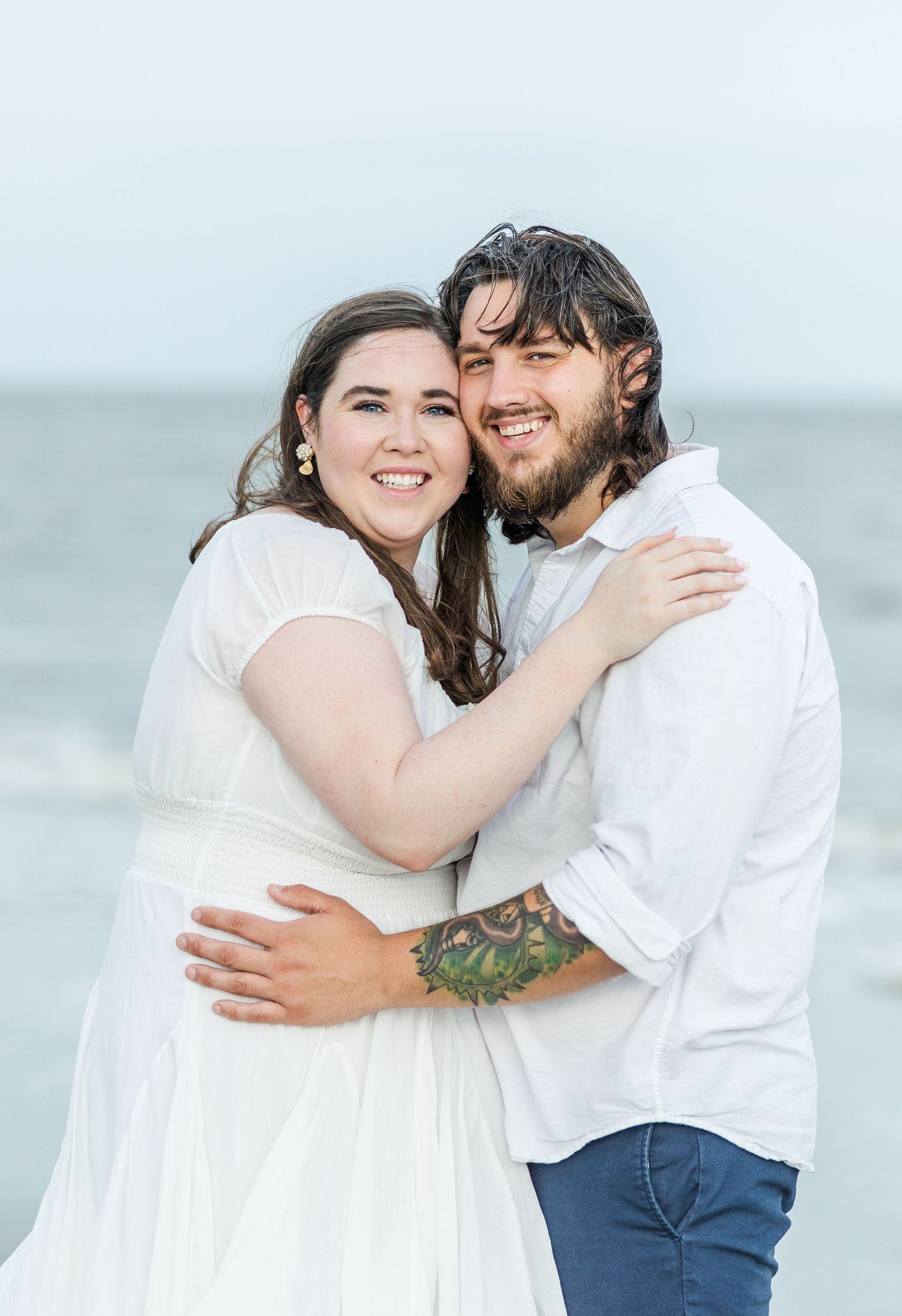 A romantic couple embraces on the beach wearing white clothing, with the ocean visible in the background.