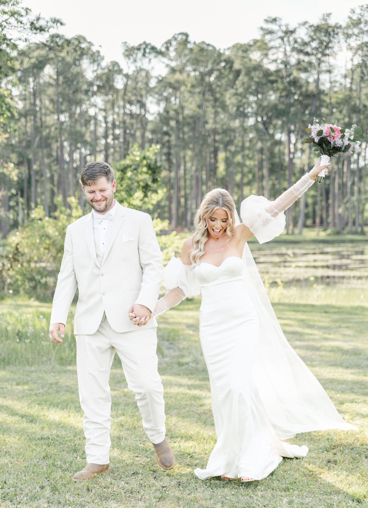A joyful wedding couple in all-white outfits celebrate on a grassy lawn with tall pine trees in the background.
