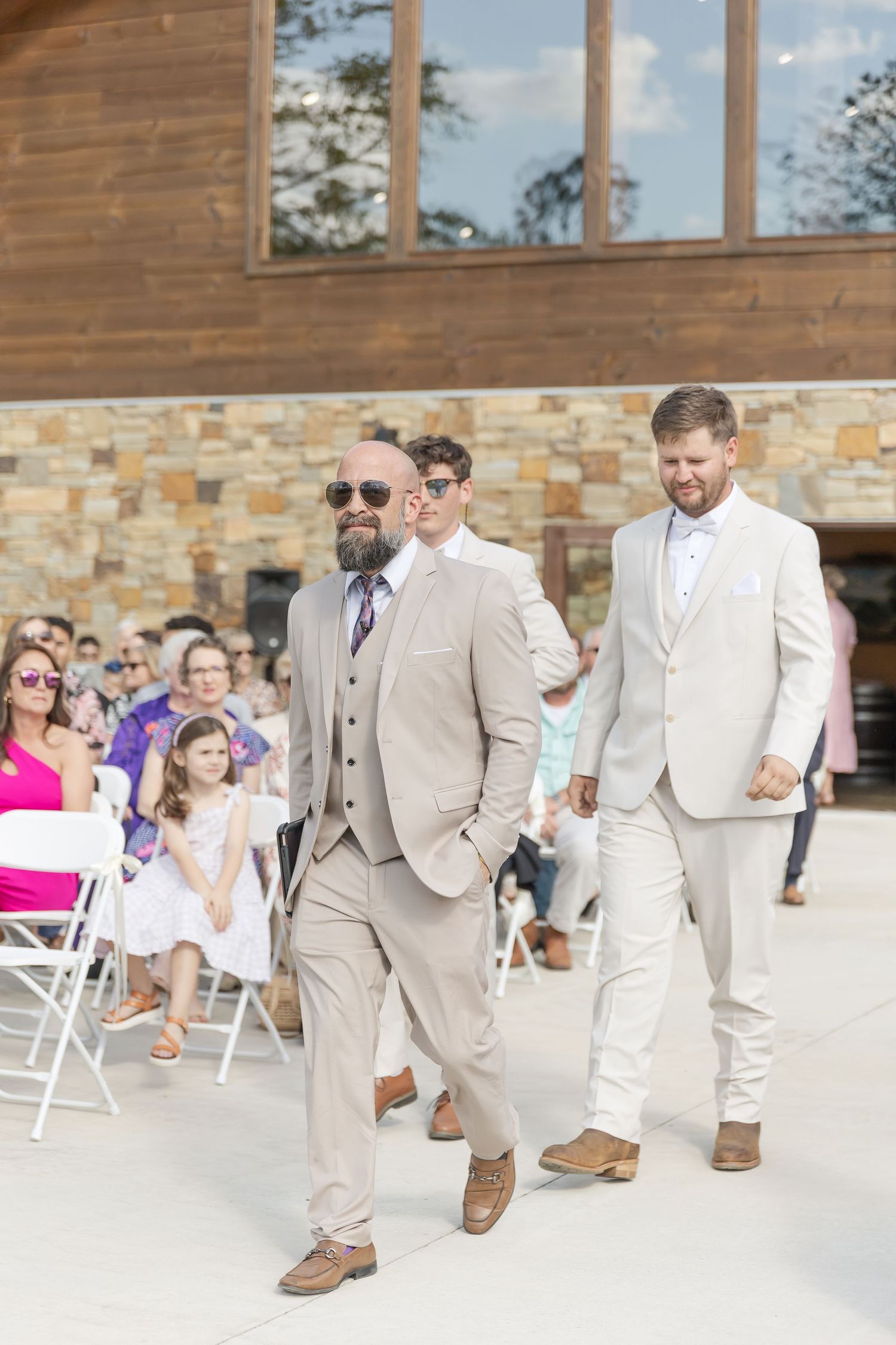 Three groomsmen in coordinating beige suits walk past wedding guests seated in white chairs.