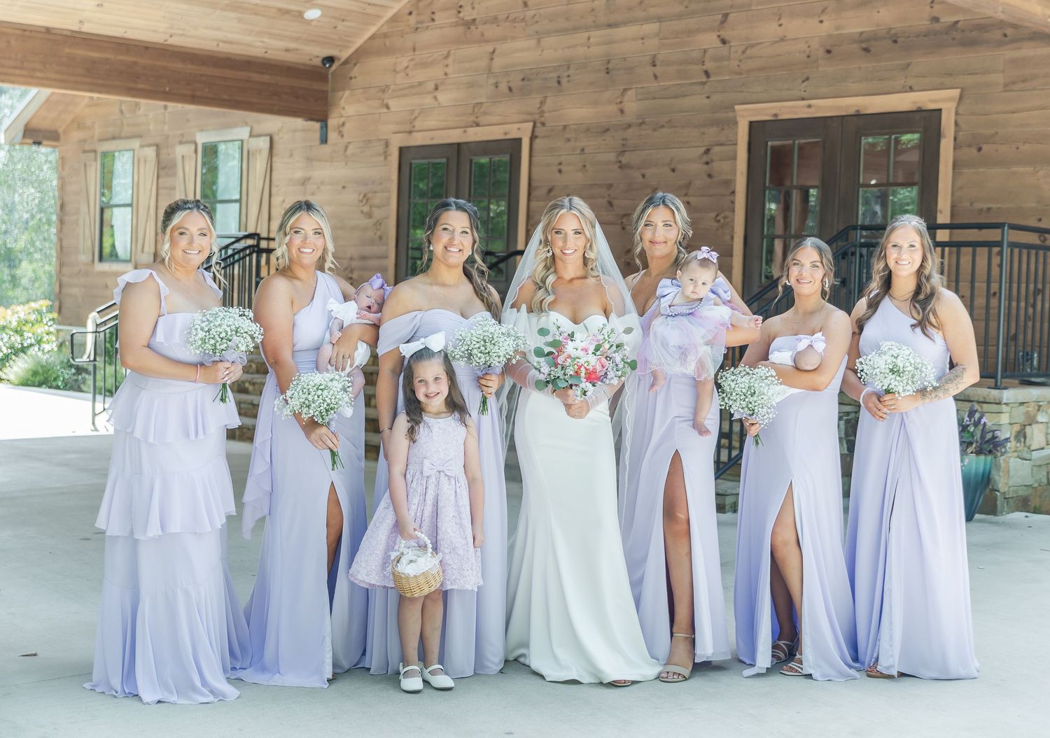 Bridal party in lavender dresses poses with bride in white gown holding pink and white bouquets.
