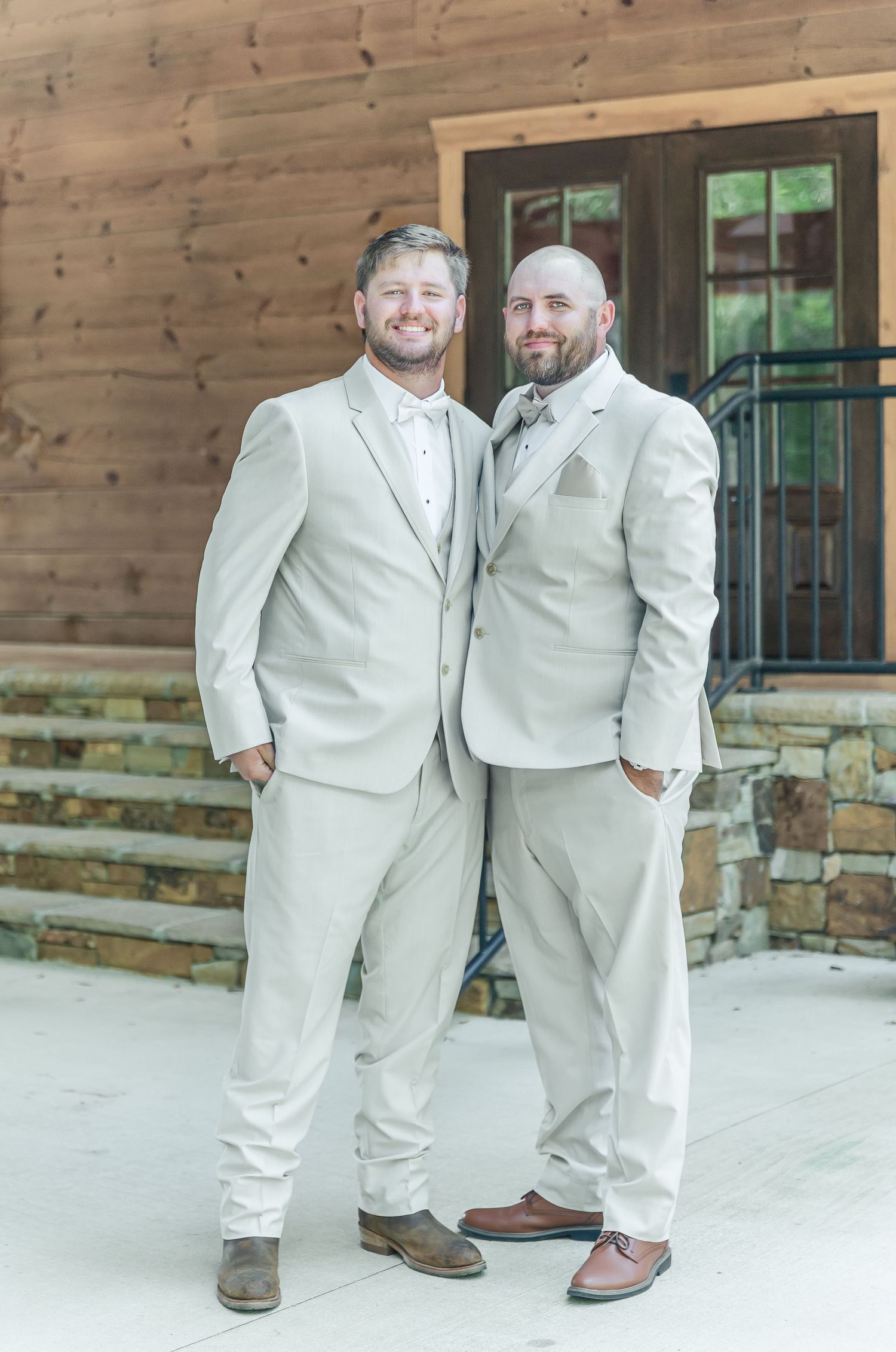 Two people wearing light beige suits pose together in front of a rustic wooden cabin exterior.
