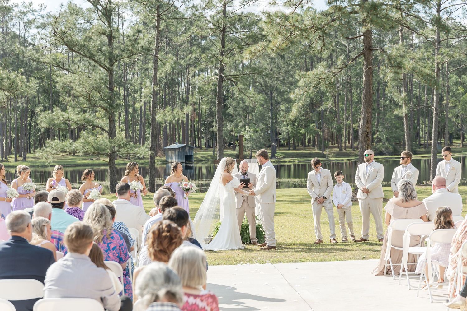 A scenic outdoor wedding ceremony with guests seated on white chairs and tall pine trees in the background.