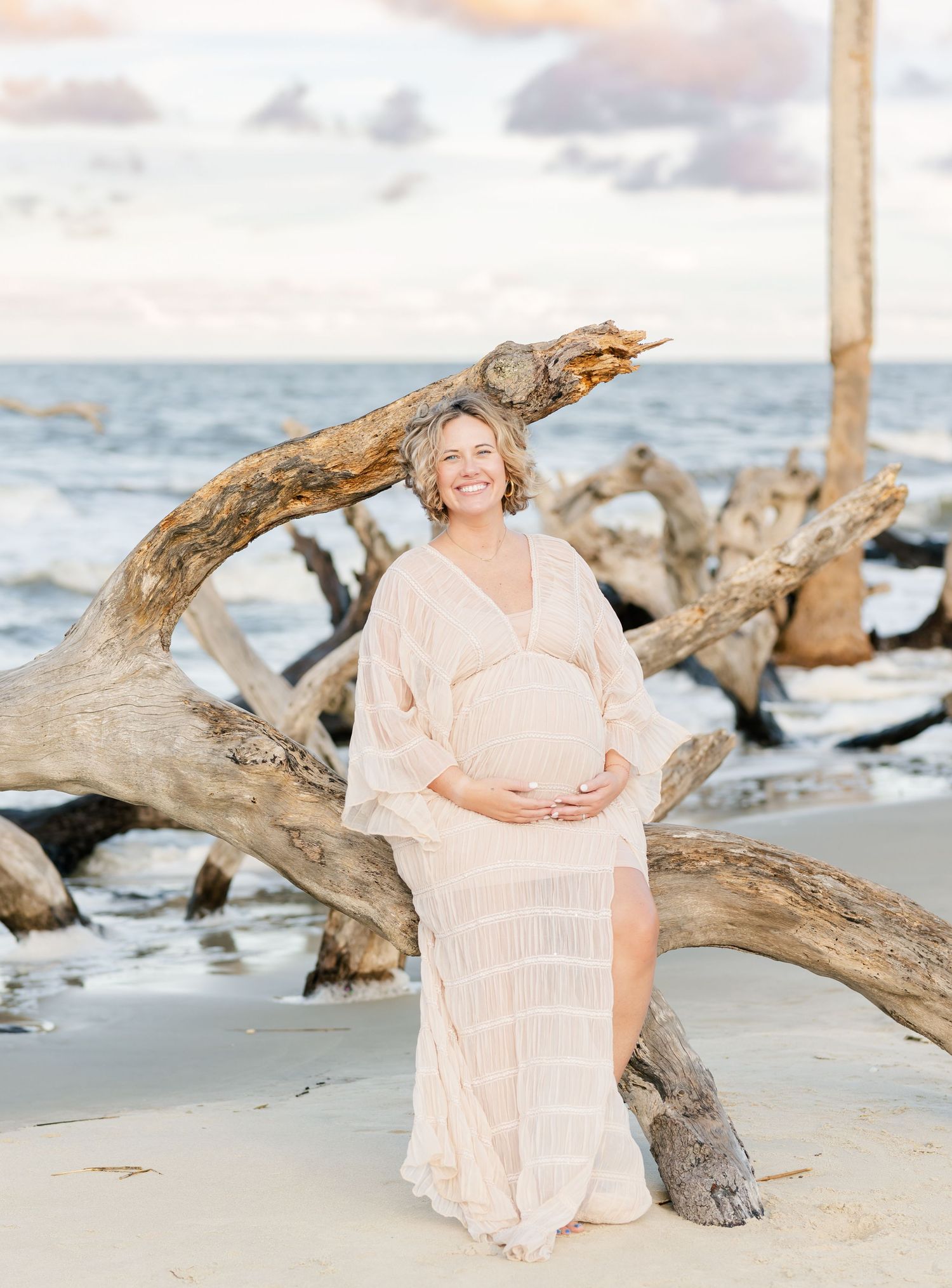 Maternity photo shoot on beach with person in light flowy dress posing next to large driftwood.