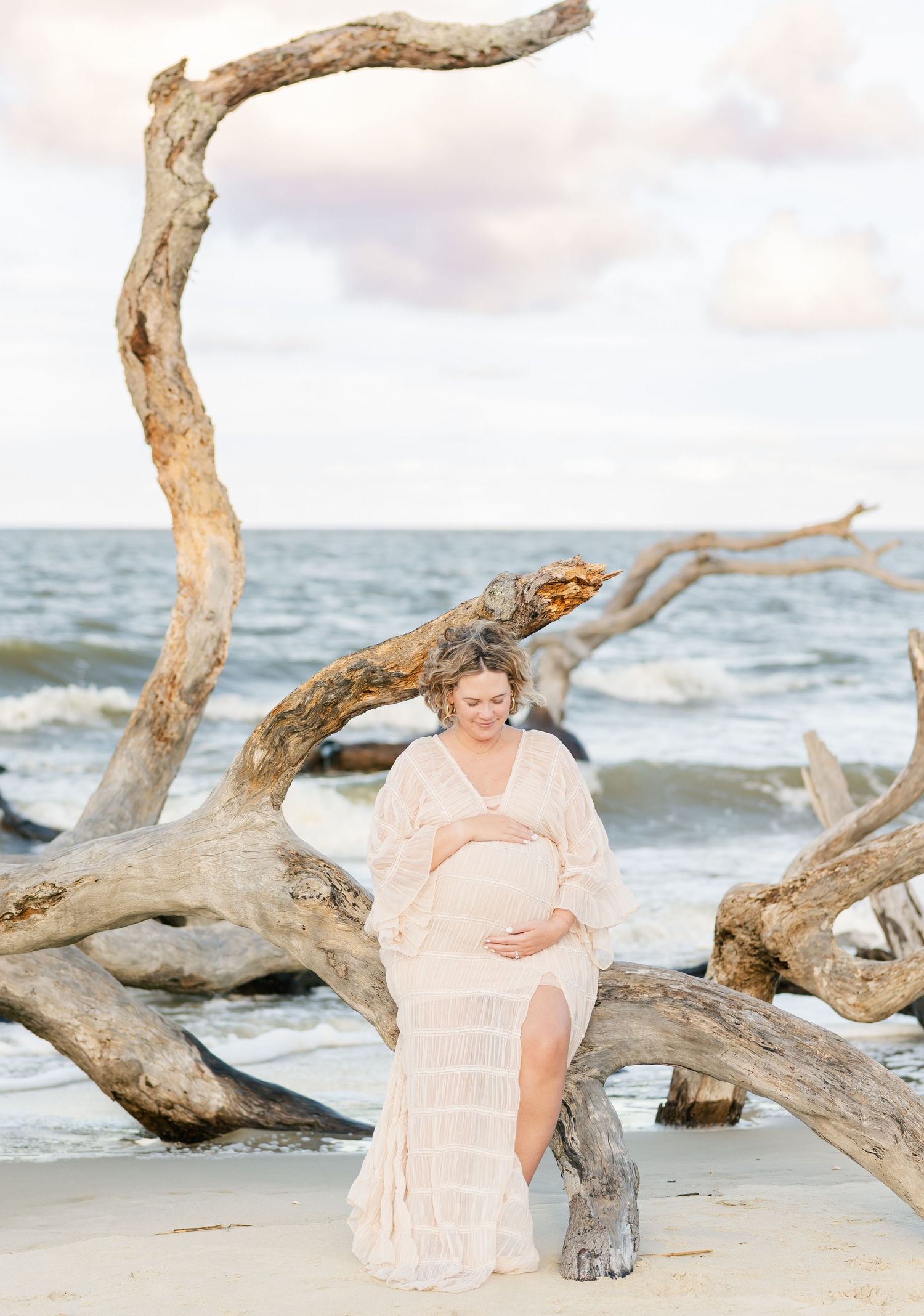 Beach maternity photography session captures a figure in a soft peach dress among natural driftwood at the ocean's edge.