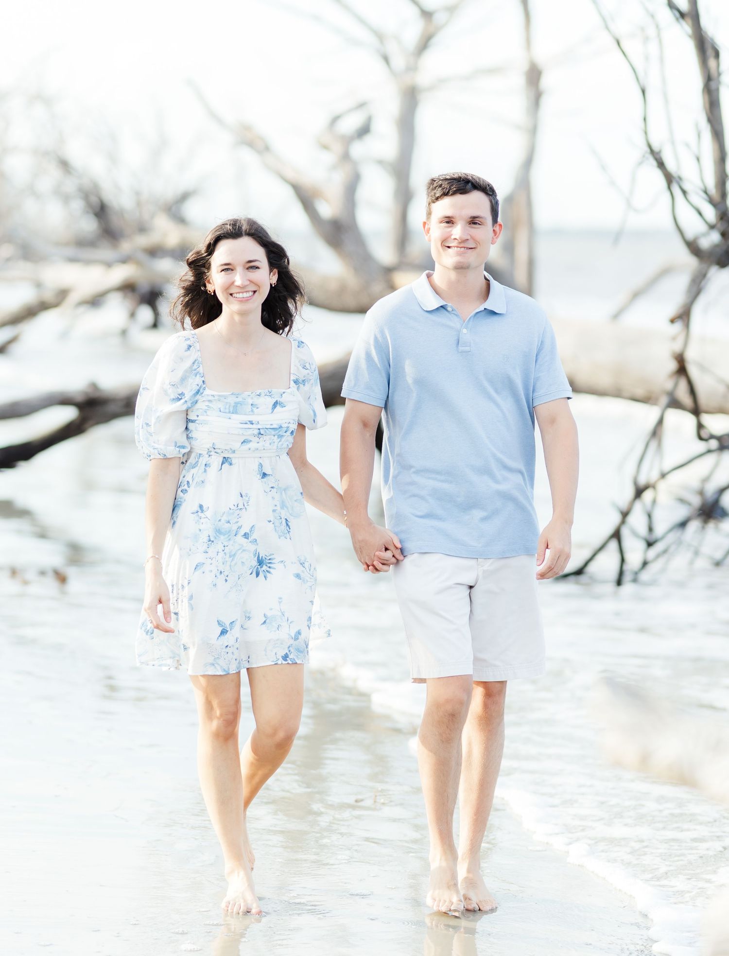 A couple walks hand in hand along the shoreline wearing matching blue and white outfits.