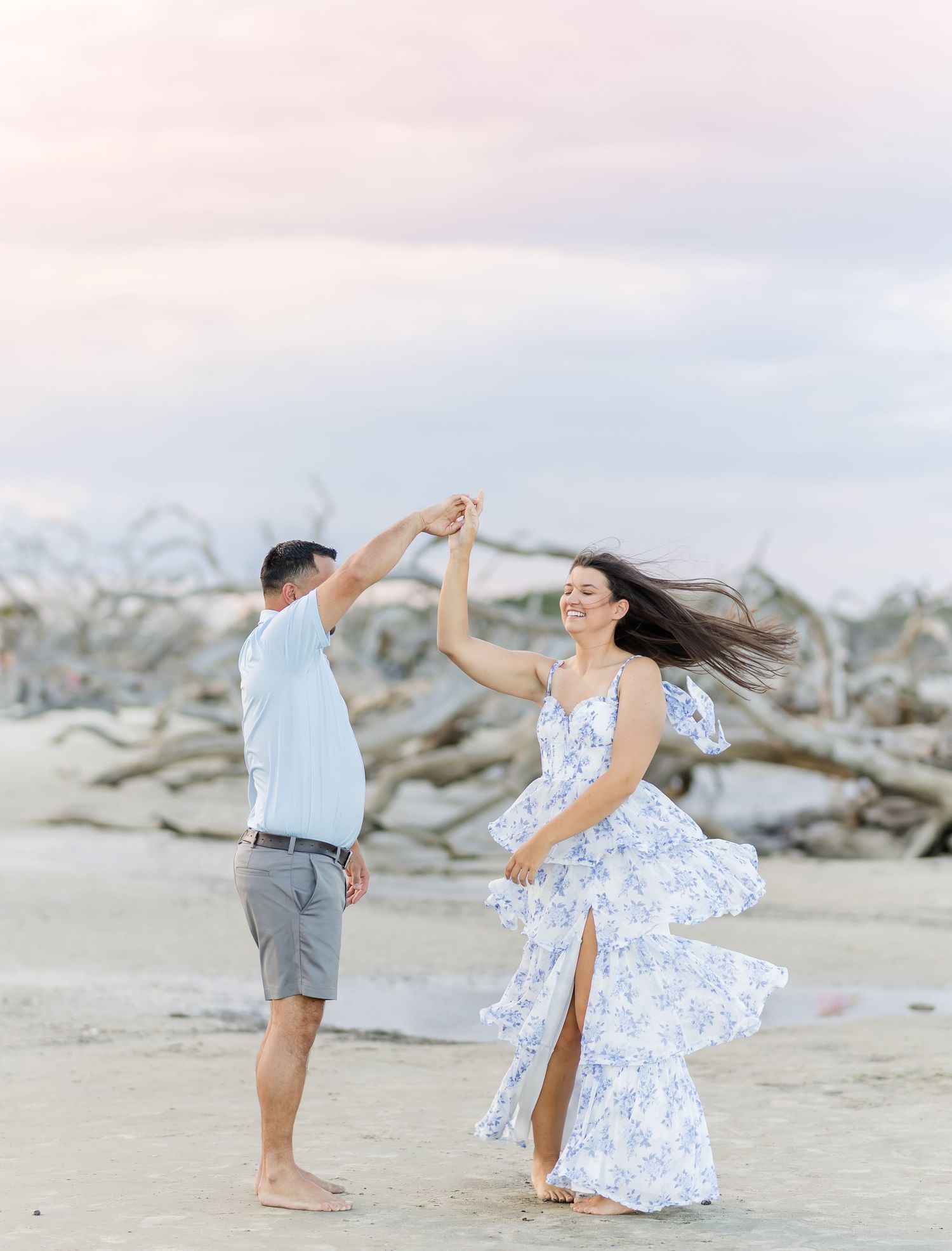 A couple dances playfully on a scenic driftwood beach at sunset with flowing dress and casual attire.