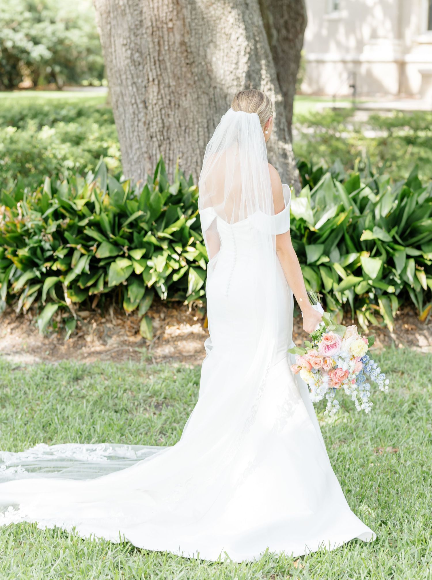 A flowing white wedding dress train spreads beautifully across green grass and garden foliage.