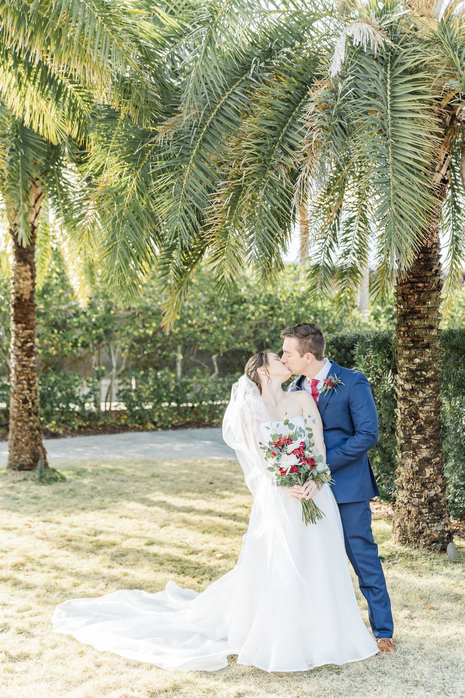 A romantic wedding portrait under palm trees with a couple sharing a kiss while holding a red bouquet.