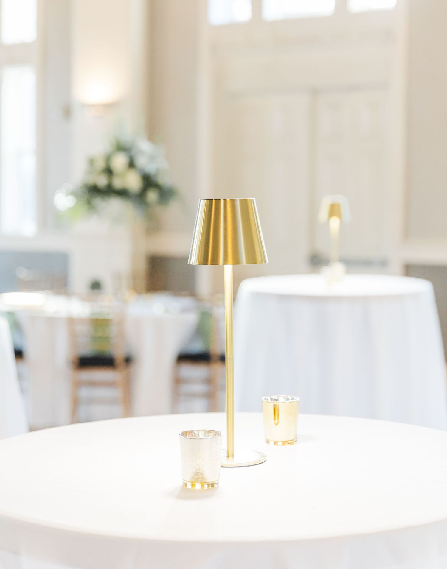 Gold cordless table lamp with votive candles on white linen cocktail table in bright wedding venue.