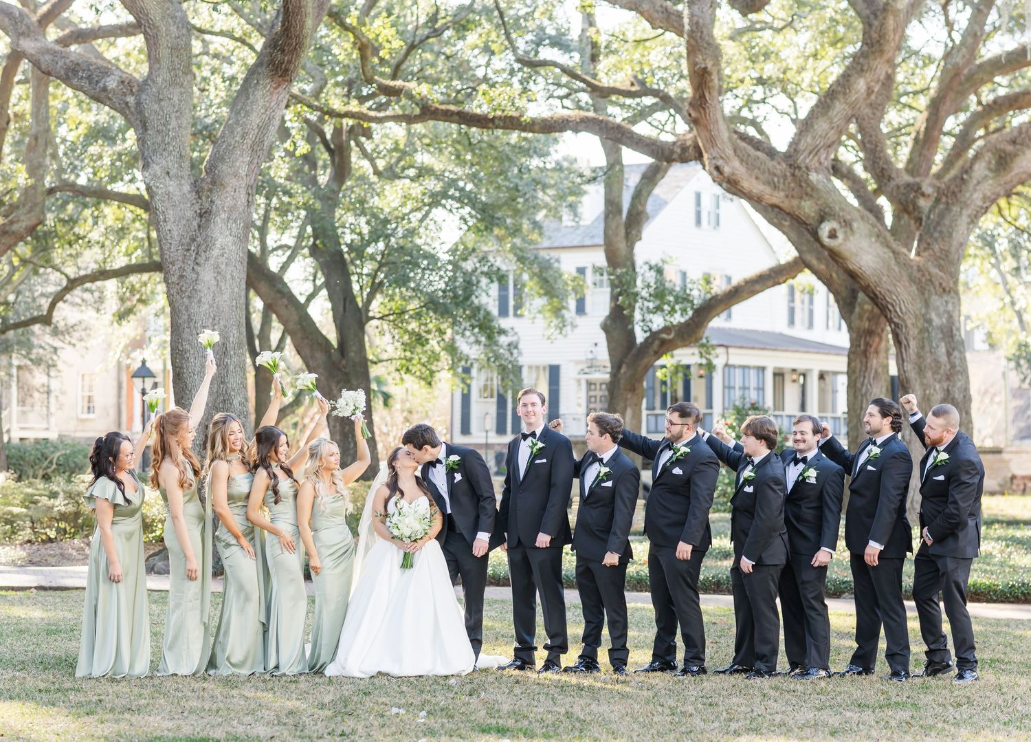 Bride and groom share a kiss surrounded by wedding party in sage green and black formal attire under oak trees.
